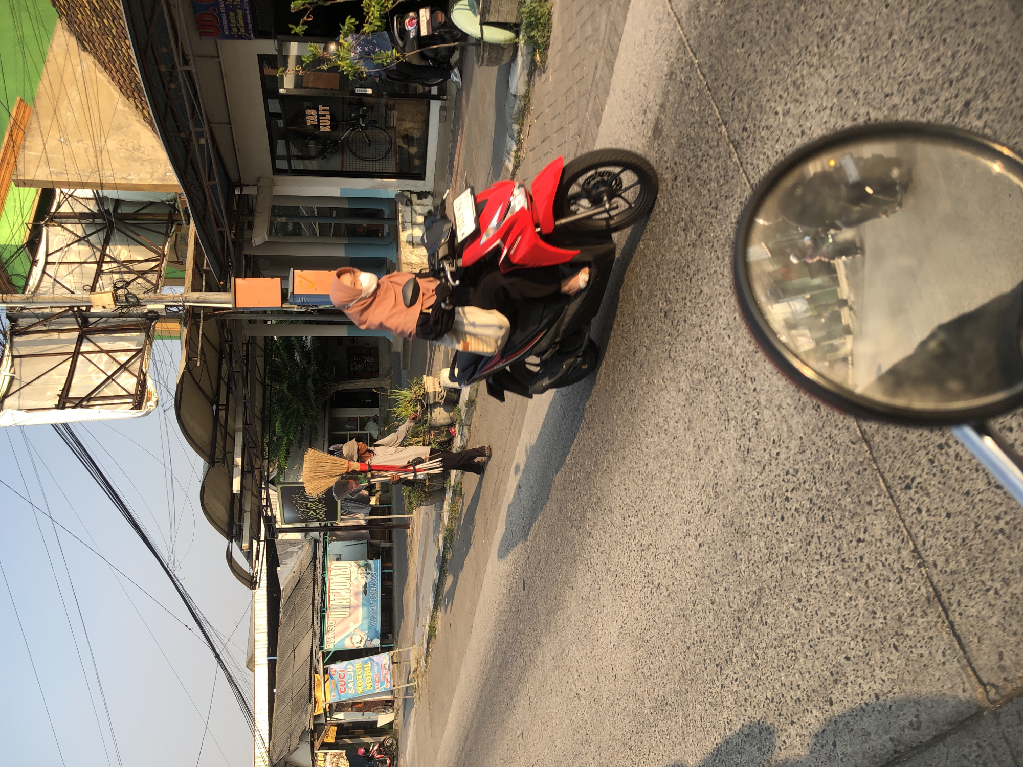 This image captures a street scene in Boro, Indonesia, likely during the golden hour of either sunrise or sunset, indicated by the warm, long shadows cast across the asphalt. In the foreground, a red motorcycle is in motion, viewed from the perspective of another rider, as evidenced by the motorcycle's rearview mirror reflecting a blurry scene of traffic.
On the right side of the street, a woman wearing a hijab and a face mask rides a red motorcycle, also in motion. She is wearing a light brown hijab and a white face mask, and a beige bag is slung over her shoulder. Next to her, a person dressed in a light-colored long-sleeved shirt and dark pants is walking and carrying a bundle of brooms and brushes over their shoulder. This individual appears to be a street cleaner or vendor.
The background features a row of buildings with awnings and signs, suggesting a commercial area. One sign clearly visible reads "TAS KULIT," which translates to "Leather Bag." Another sign advertises "BAKSO URAT JUMBO" (Jumbo Vein Meatball). Several other signs are present, though their text is less legible. Overhead, a complex network of power lines crisscrosses the sky. The street is paved with asphalt and bordered by a sidewalk with potted plants. The overall atmosphere is that of a typical urban environment in Indonesia, with the presence of motorcycles, street vendors, and commercial establishments.