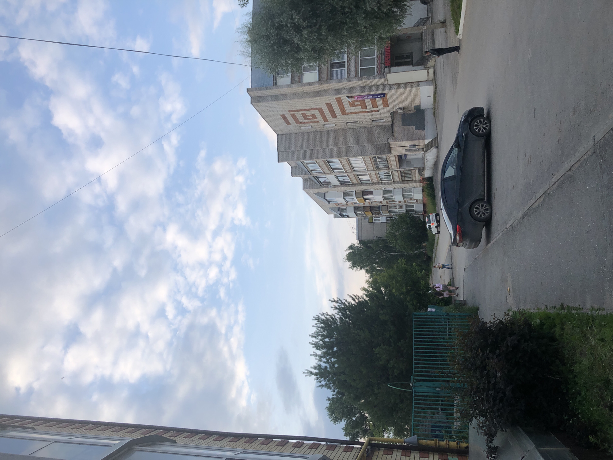 The image shows a street scene in Solikamsk, Russia, likely during the daytime given the bright sky with scattered clouds. A multi-story brick building dominates the background, featuring a large geometric pattern on its facade. In front of the building, a dark-colored car is parked on the asphalt street. A sidewalk runs alongside the street, and several people are visible walking and standing on it. A green metal fence and some trees and bushes are also present in the foreground. The overall atmosphere appears to be that of a typical urban residential area. There is some visible text on the building, but it is not clearly legible.