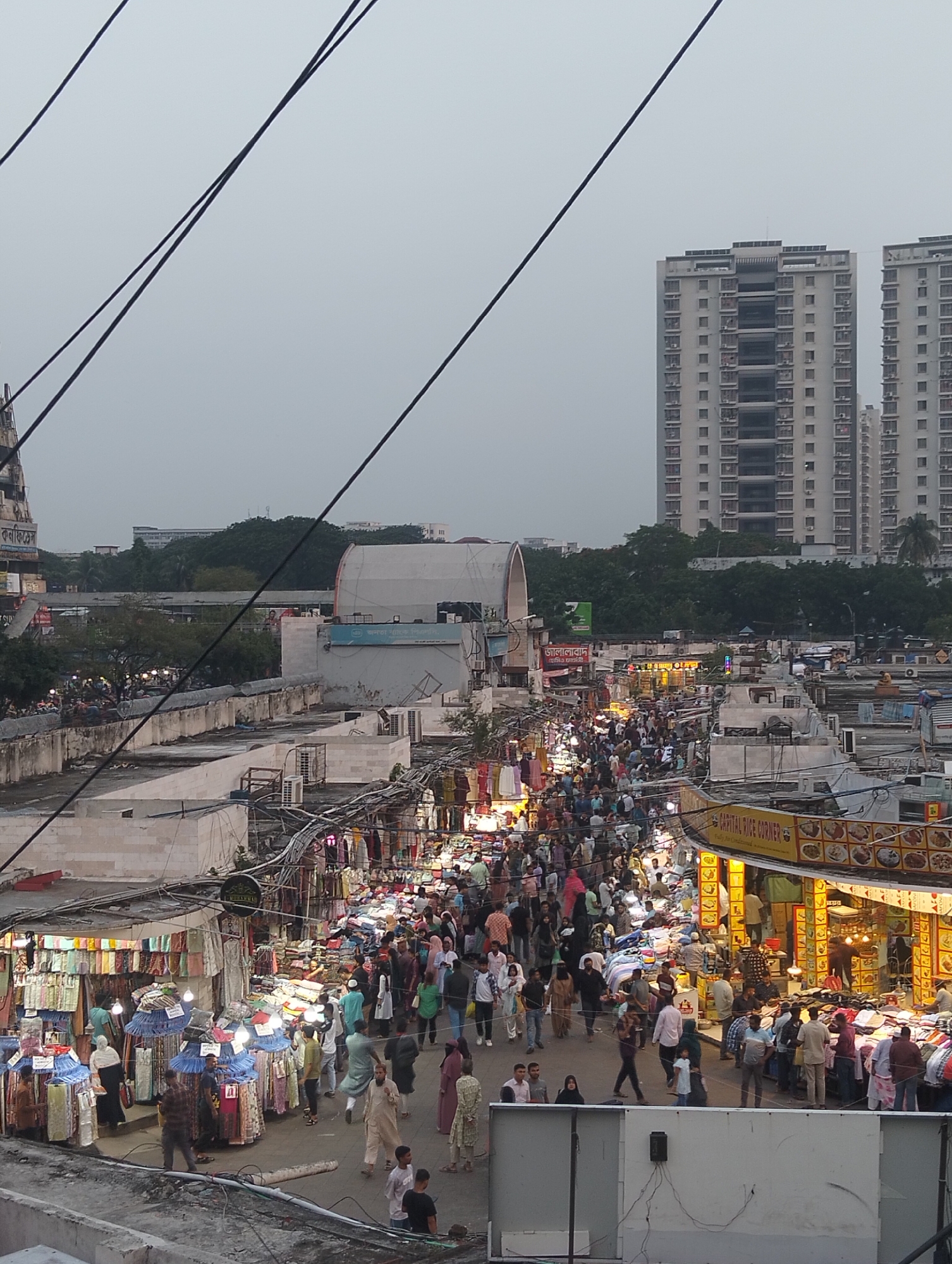 The image captures a bustling street scene in Dhaka, Bangladesh, likely during an evening. The area is crowded with people, suggesting a market or shopping district. Numerous stalls and shops line the street, illuminated by artificial lights. The stalls sell clothing, fabrics, and food. Many individuals are browsing the merchandise or walking through the crowded street. Above the stalls are a tangle of wires and cables. In the background, tall residential buildings rise into the hazy sky, typical of Dhaka's urban landscape.