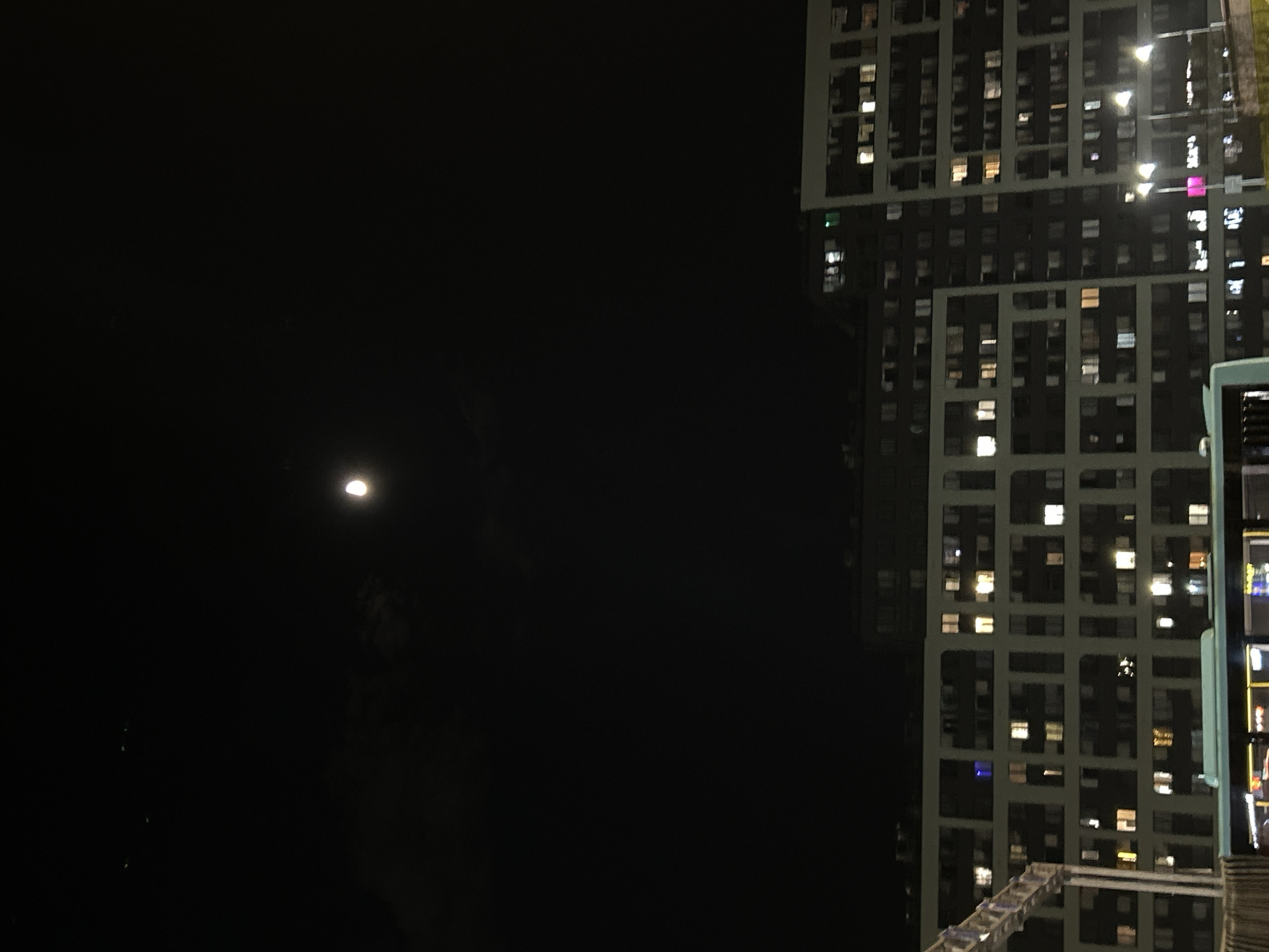 The image shows a nighttime view in Saint Petersburg, Russia, dominated by a tall, modern apartment building with numerous illuminated windows. The building’s architecture features a grid-like pattern formed by the window arrangement and structural elements. A smaller platform or roof structure is visible at the base of the building, possibly part of an adjacent structure. To the left of the building, the scene fades into darkness, but a faint moon is discernible in the night sky. The overall ambiance is urban and nocturnal.