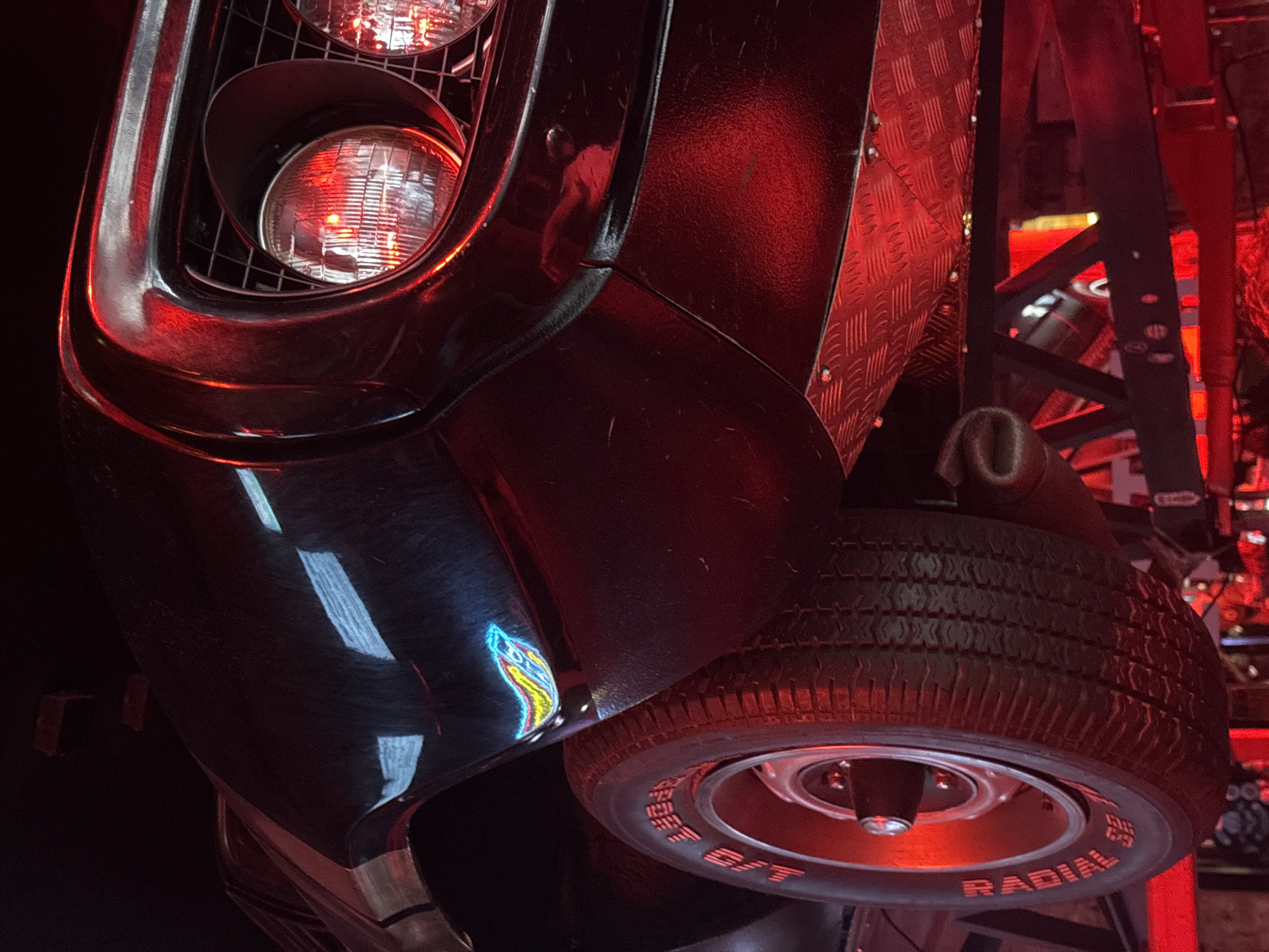 The image shows the front of a black vehicle, possibly a classic car, in a location with red lighting. The vehicle has visible headlights and a tire with "RADIAL" and "STREET B/F" markings. Reflections on the glossy black paint suggest the presence of other objects or light sources in the vicinity. The background includes metal grating and structural elements, which may indicate an industrial or entertainment setting. Without additional context, it's not possible to confirm the location in Saint Petersburg, Russia, but the scene suggests a curated or stylized environment.