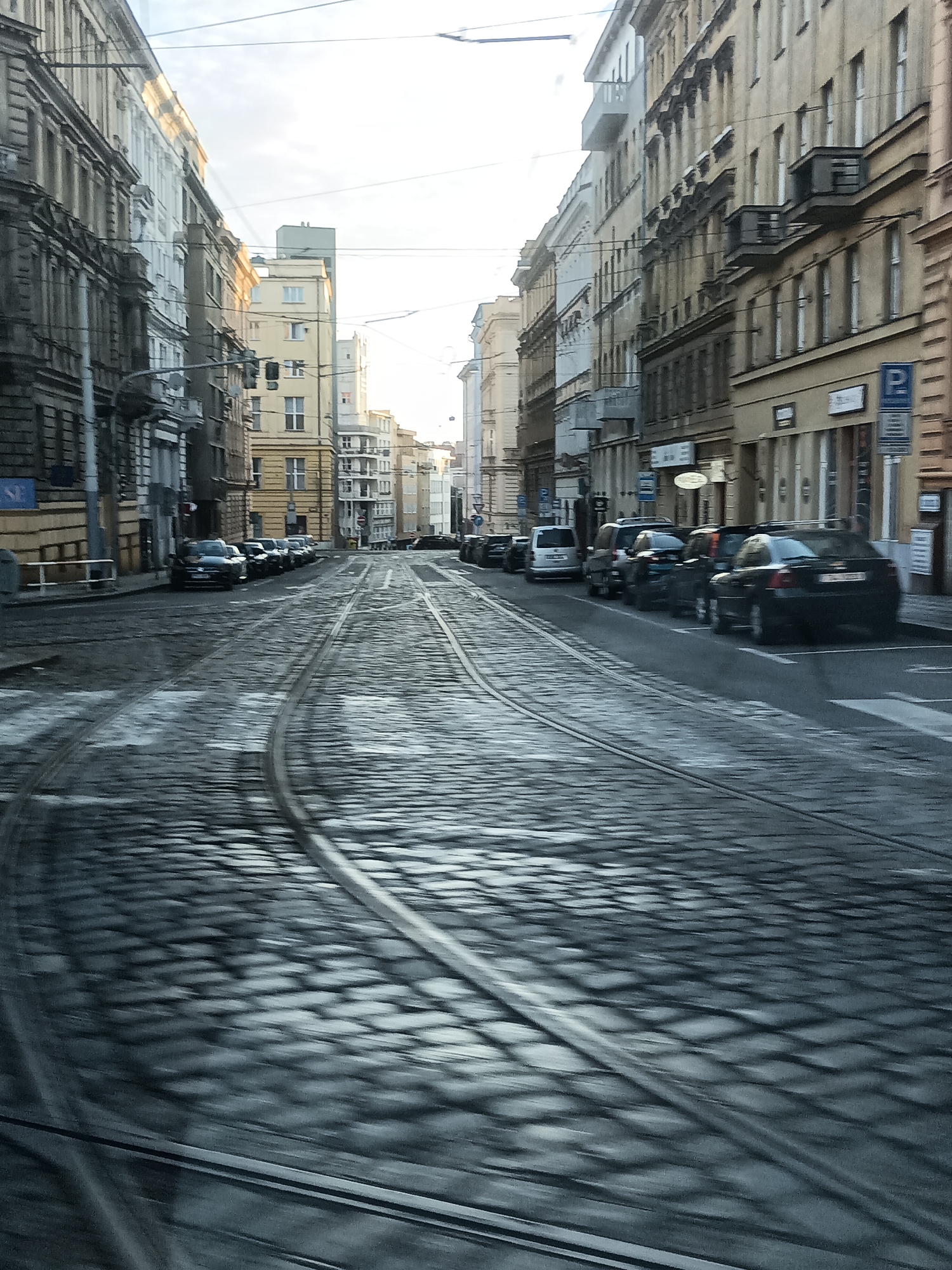 The image depicts a street scene in Prague, Czechia, characterized by cobblestone paving and tram tracks. The perspective suggests the photograph was taken from a moving vehicle, possibly a tram, given the motion blur. Lined with multi-story buildings, the street appears to be a commercial or residential area. Several cars are parked along the right side of the street, and there is a visible blue parking sign. The sky is bright and overcast. The overall impression is that of a typical European urban environment during the day.