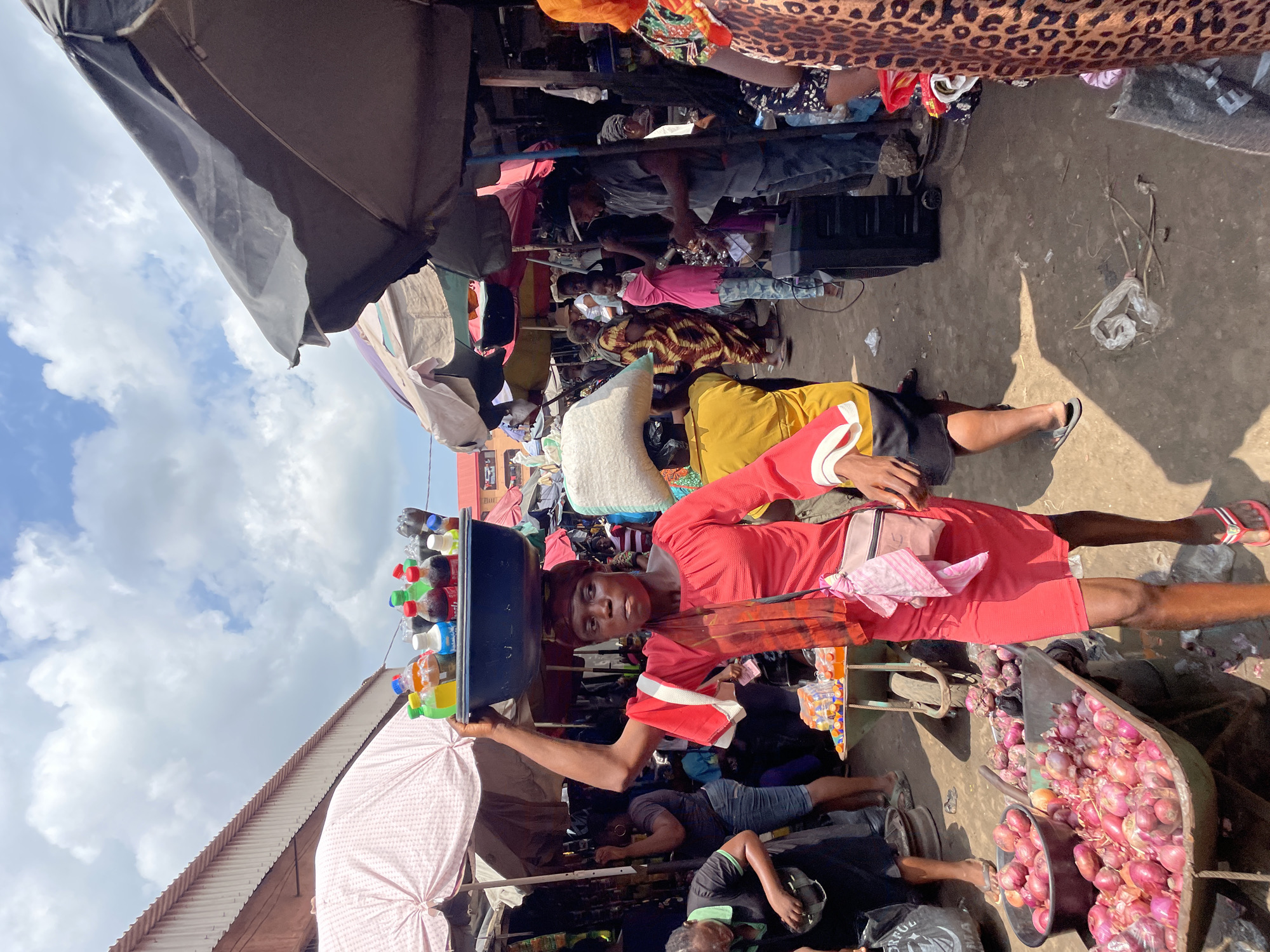 This full-shot image captures a vibrant and bustling outdoor market scene in Fegge, Nigeria, under a bright, sunny sky with scattered white clouds. The ground is a mix of dirt and uneven concrete, with some litter like plastic bags and sticks visible.
In the immediate foreground, a woman in a long-sleeved red dress and red sandals is the central focus. She carries a large, dark blue basin filled with various bottled beverages (sodas, water, and juices in different colored plastic bottles) balanced expertly on her head. Her right arm is raised, supporting the basin, while her left arm hangs down. She wears a small, light-colored purse slung across her body and a patterned scarf or fabric around her neck. Her gaze is directed straight at the viewer, with a neutral expression.
To the woman's right, a large, dark-colored cart overflows with numerous red onions, some contained in a smaller black bowl within the cart. Another person is partially visible near the cart.
The background and mid-ground are populated with many other individuals, suggesting a crowded market environment. People are seen standing, sitting, and crouching, some under the shade of various market structures. One person is wearing a distinctive leopard-print fabric. Market stalls, constructed from simple frames and covered with tarps or umbrellas in dark grey/black and lighter pink/white, line the area, providing shade from the sun. A large white sack, likely containing dry goods, is also visible. The activity appears to be the daily commerce of a street market.
The strong sunlight creates distinct shadows on the ground. No discernible text is clearly visible in the image.