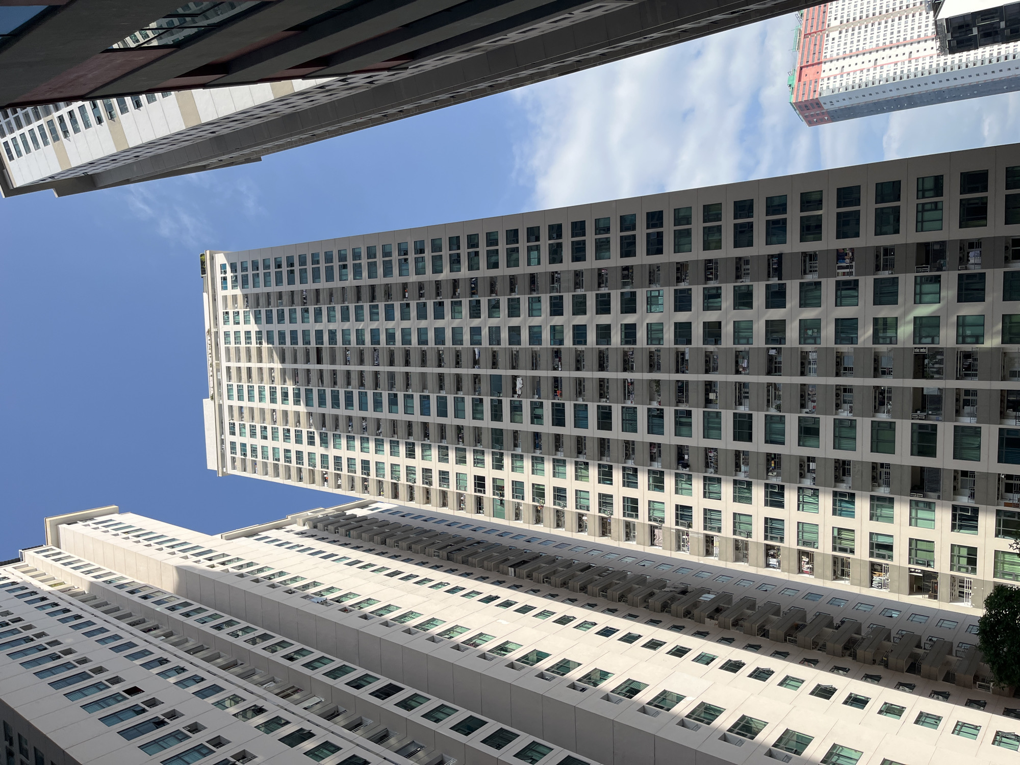The image depicts a low-angle, upward-looking view of several high-rise residential buildings under a bright blue sky with scattered white clouds, located in Bac Nha Trang Ward, Vietnam.
In the center, a prominent, light beige-colored high-rise apartment building dominates the frame, extending from the bottom almost to the top. It features numerous uniformly spaced, green-tinted windows and balconies on its main facade, with clothes hanging on several balconies, indicating habitation. The building's front is brightly lit by direct sunlight, casting distinct shadows on its recessed architectural features and also onto the building immediately to its left.
To the left, a portion of another similar light-colored high-rise building is visible, aligning vertically with the central structure and exhibiting comparable window and balcony patterns.
To the immediate right, a dark grey or brownish building edge is visible, providing a narrow, vertical frame on that side and partially obscuring the sky.
In the far background, on the right side, another very tall residential building with a distinctive red and white/light grey color scheme is visible. This building appears to be of a different architectural style or a newer construction.
A small patch of green foliage is visible at the very bottom left corner, indicating the presence of ground-level vegetation. No people are visibly depicted in the frame. The strong sunlight and clear sky suggest a daytime scene.
