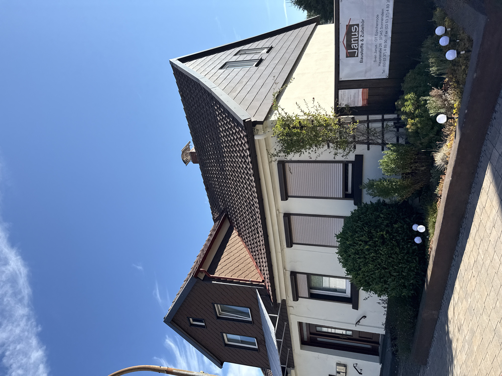 The image shows the exterior of a two-story building with a pitched roof, located in Walkenried, Germany. The building has a white facade on the lower level and brown shingle-like siding on the upper level and parts of the roof. There are several windows, some with closed roller shutters. A dark brown door is visible on the left side of the building. A sign for "Janus Baustoffe & Zubehör" is mounted on a dark fence in front of the building, displaying contact information. The foreground features a paved walkway and a garden bed with various plants and several spherical outdoor lights. The sky is bright blue with scattered white clouds. There are no people visible in the image.