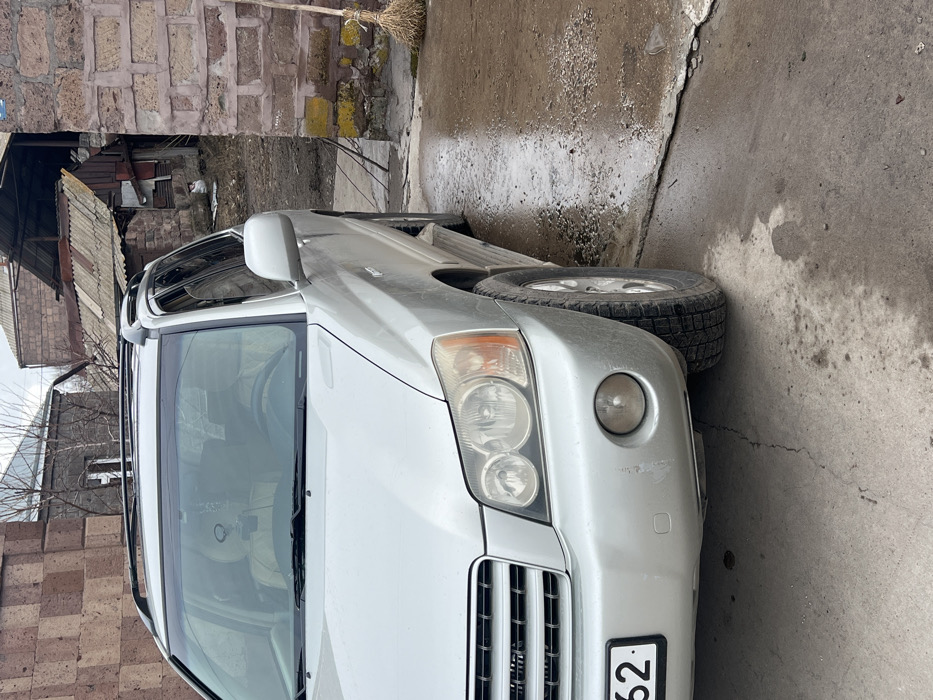 The image depicts the front and driver's side of a silver SUV, consistent with a Toyota Highlander model, parked on a concrete surface in Azatan, Armenia. The vehicle's front bumper, grille, and headlight assembly are prominent. The front bumper exhibits minor scuffs on the driver's side and appears slightly misaligned below the grille. The headlights show some hazing. A partial license plate ending in "62" is visible on the lower left of the front bumper. Roof rails are present on the vehicle's roof, and side steps are visible below the driver's door. Through the windshield, light-colored seats and a dark object hanging from the rearview mirror are discernible.
The concrete ground beneath and immediately to the right of the vehicle is wet, showing puddles and accumulated dirt/grime. A brick wall with patches of yellow-green discoloration runs along the right side of the frame, against which a natural-fiber broom leans. Beyond this wall, a dirt path leads to several dilapidated structures with weathered corrugated metal roofs and brick/stone walls. A small white object rests on the ground near these structures. In the background to the left, behind the vehicle, are other buildings constructed from red brick and stone under an overcast sky. No individuals are present, and no activity is occurring in the scene.