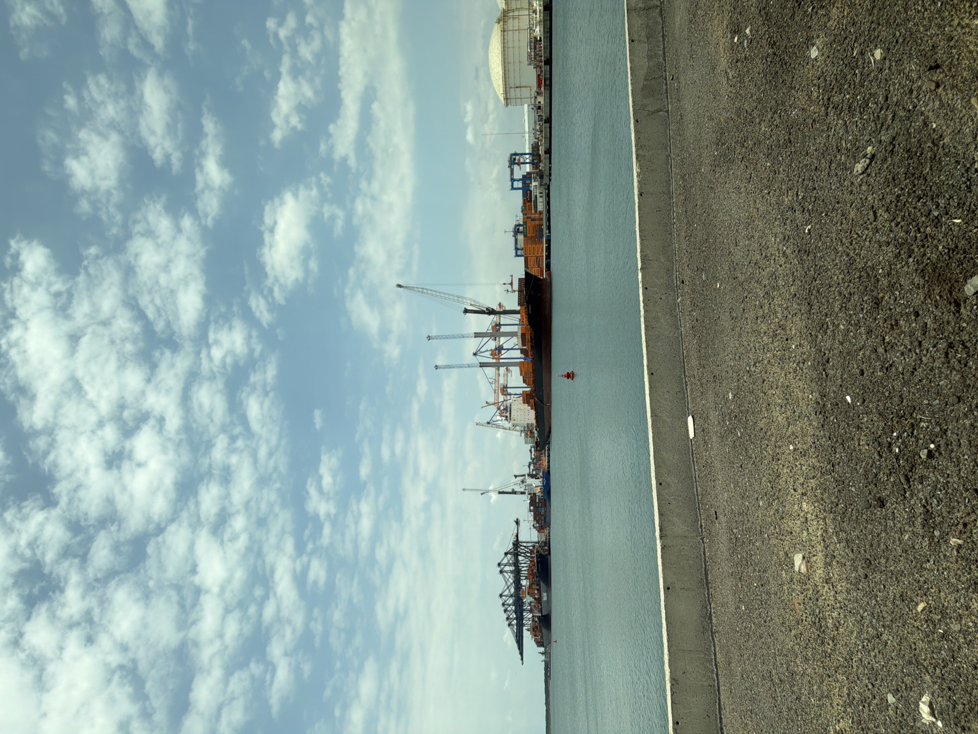 The image shows a port in Veracruz City, Mexico. Several large cargo ships are docked along a concrete quay. Cranes are visible on the ships and along the dockside. Behind the ships, large industrial buildings, possibly storage tanks, can be seen. The sky is blue with scattered white clouds. A concrete edge separates the port from a gray, gravelly surface.