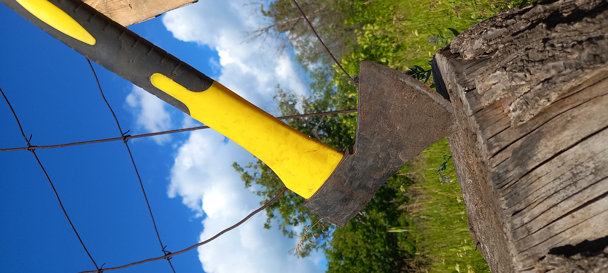 The image depicts an axe with a black and yellow handle embedded in a weathered wooden log. A wire fence crosses the view, with a blue sky dotted with clouds visible in the background. Green vegetation is also visible in the backdrop.