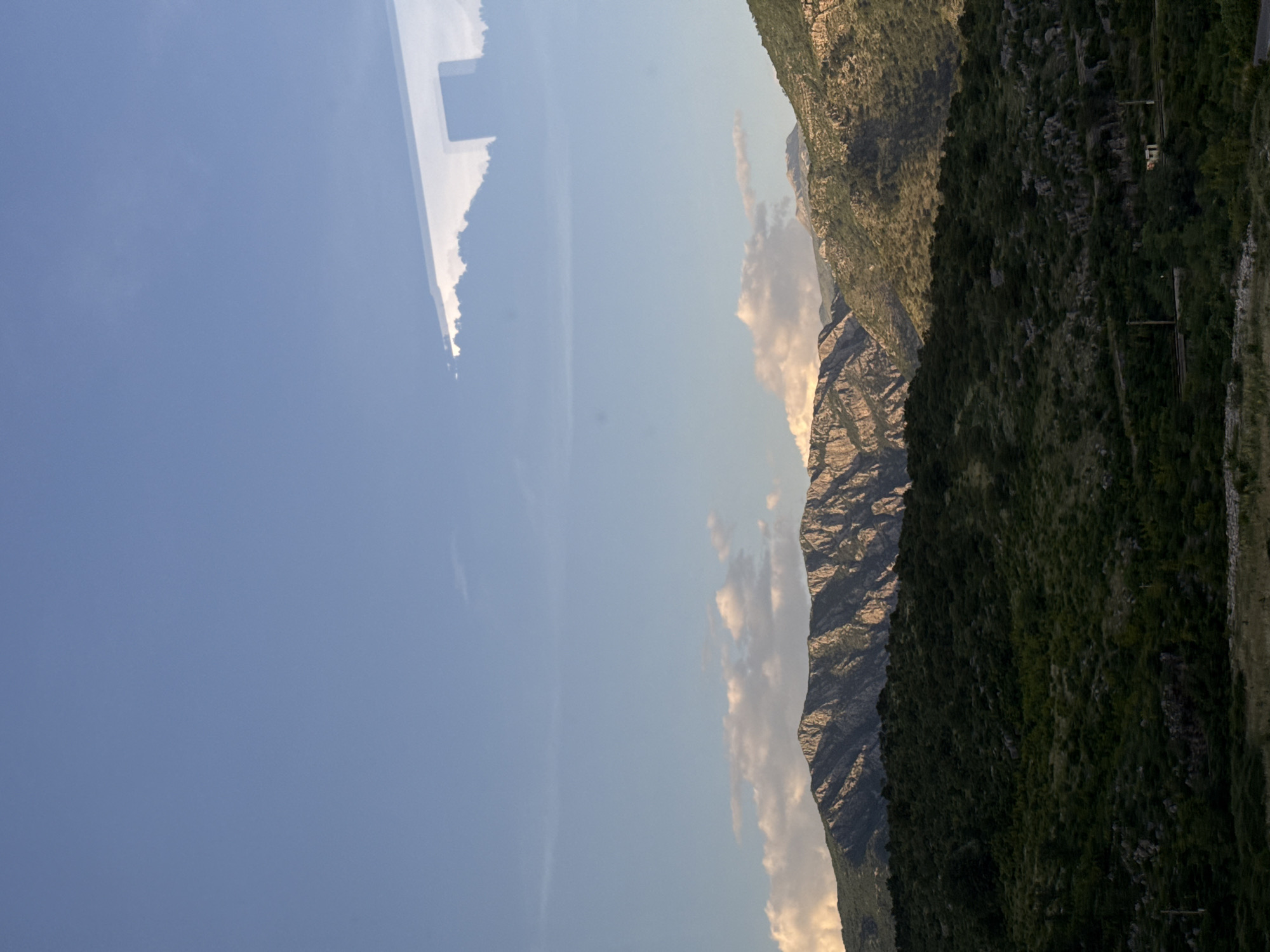 The image shows a landscape in Klopot, Montenegro, featuring a clear blue sky with some wispy clouds on the left. On the right side of the image are mountains, with exposed rock faces and green vegetation covering some areas. There is also a road running along the right side of the mountain, with telephone poles visible. The light suggests it could be either sunrise or sunset.