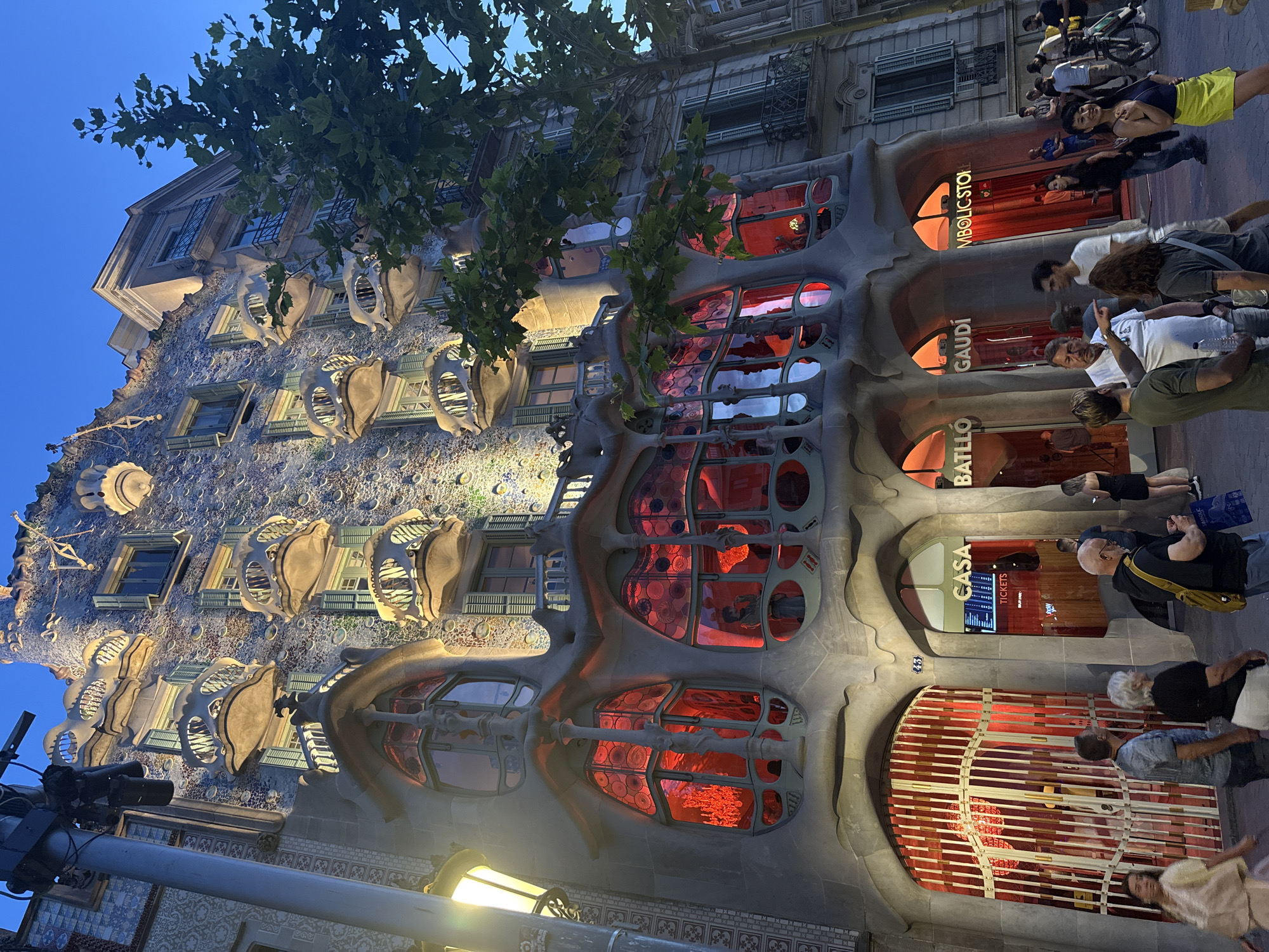 The image shows Casa Batlló, a building designed by Antoni Gaudí, located on Passeig de Gràcia in Barcelona, Spain. The building's facade is characterized by its organic, undulating forms and mosaic-like surface, with distinctive balconies that resemble skulls and bones. The large, rounded windows on the lower floors are illuminated with a vibrant red light. In the foreground, a group of people are standing on the sidewalk, some looking at the building, while others are engaged in conversation. A street lamp is visible to the left of the building. The sky above is a deep blue, indicating it is either dusk or early evening. The overall scene captures the unique architectural style of Gaudí and the lively atmosphere of the city.
