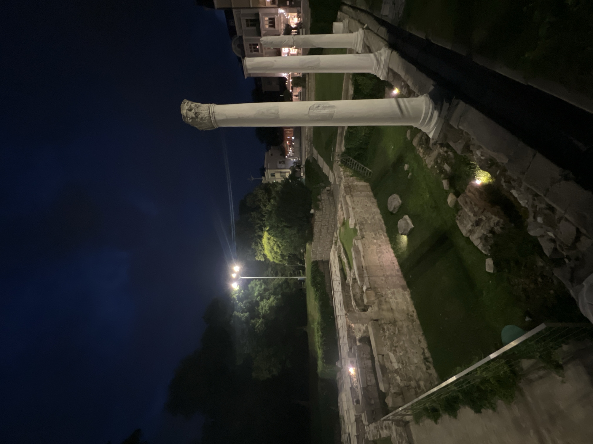 The image depicts the Roman Forum in Plovdiv, Bulgaria, at night. The scene is illuminated by artificial lighting, casting long shadows and highlighting the ancient ruins. In the foreground, several tall, white marble columns stand against a dark blue night sky. The tops of these columns are adorned with ornate capitals. To the left of the columns, the remains of stone structures, likely part of the ancient forum, are visible. These ruins are low-lying and partially covered by grass. The area is sectioned off by a metal fence with greenery growing along it. In the background, trees and the facades of buildings can be seen, also illuminated by artificial lights, suggesting the forum is situated within or adjacent to the modern city. The overall atmosphere is serene and historical, with the ruins appearing majestic under the night sky. There are no people visible in the image.
