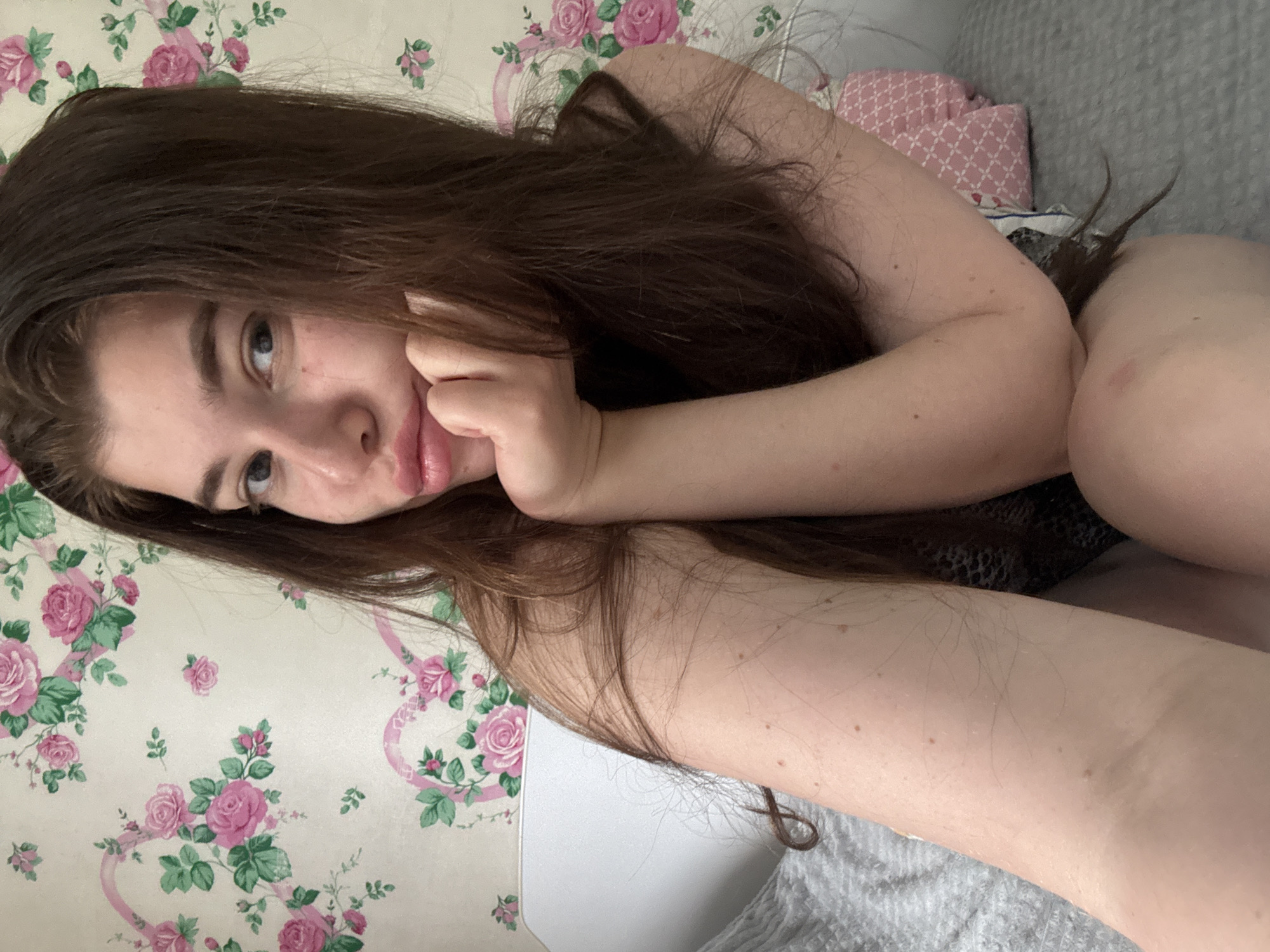 A woman with long brown hair is lying down. She is resting her cheek on her hand and looking towards the camera. The background includes a floral wallpaper and a gray patterned pillow. Location: This image was not taken in Мещёры, Russia.