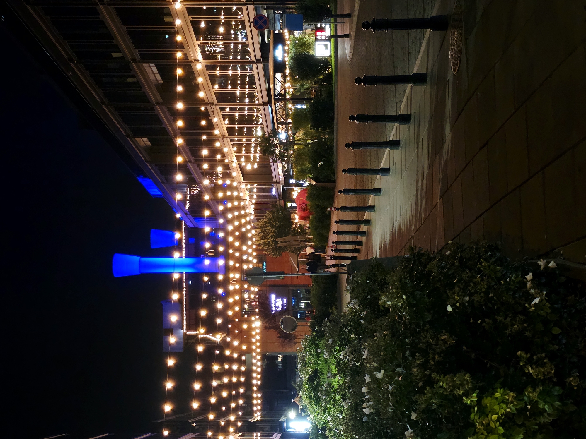 The image depicts an outdoor scene at night in Warsaw, Poland, illuminated by strings of warm-toned lights and some blue lighting. The perspective is from a low angle, looking upwards and to the right.
On the left side of the image, a modern building with a glass facade is visible, adorned with numerous hanging lights. There are also large, illuminated blue structures, possibly art installations or architectural features, against the dark night sky.
To the right of the building, a paved walkway is lined with black bollards. Beyond the walkway, there are trees and bushes, some of which are illuminated, suggesting a garden or landscaped area. In the background, further buildings and lighting can be seen, indicating an urban or commercial setting. Two figures are seated on a bench near the trees, their presence minimal in the overall scene.
The time of day is clearly night, and the weather appears to be clear, with no visible rain or fog. The ambiance is festive and inviting, likely a commercial or entertainment district due to the extensive lighting and building design. There is a traffic sign with a red circle and a diagonal bar, and another blue sign, but no other discernible text.