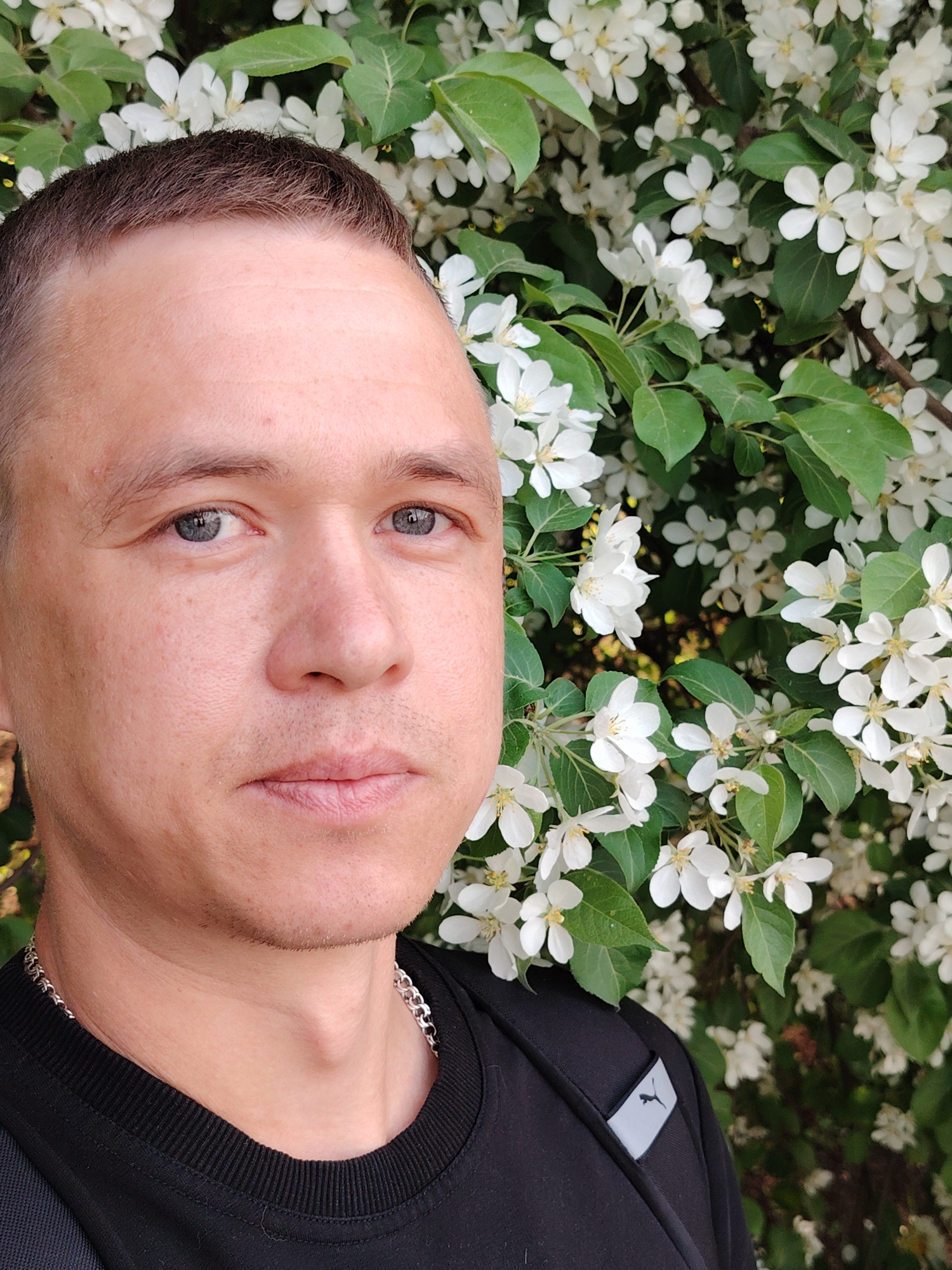 A medium shot depicts a man against a backdrop of flowering branches with green leaves and white blossoms. The man has a close-cropped haircut, light complexion, and grey eyes. He wears a black crew-neck shirt and a silver chain around his neck. A portion of a black backpack, bearing a Puma logo on a small grey tag, is visible on his left shoulder. The location is Chaikovsky, Russia, as indicated by geotag data.