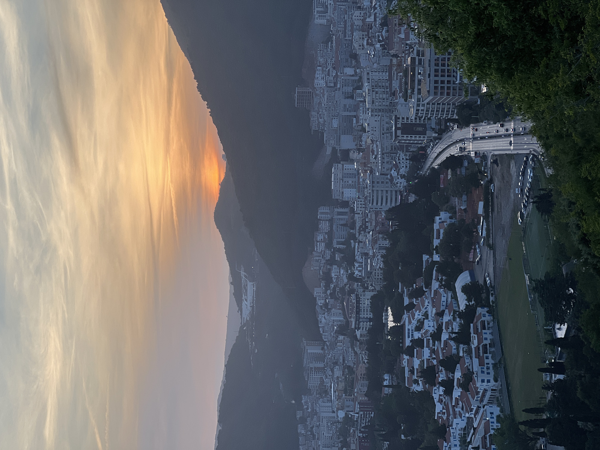 The image shows a view of the city of Budva, Montenegro, at sunset. In the foreground is a mountain range with the sun setting behind it, casting a golden glow across the sky. To the right of the mountains is the city of Budva, with its buildings and structures cascading down towards the coast. A road with moving vehicles passes through the city. Further to the right, there is a sports field. The foreground of the image shows the dark green foliage of nearby trees.