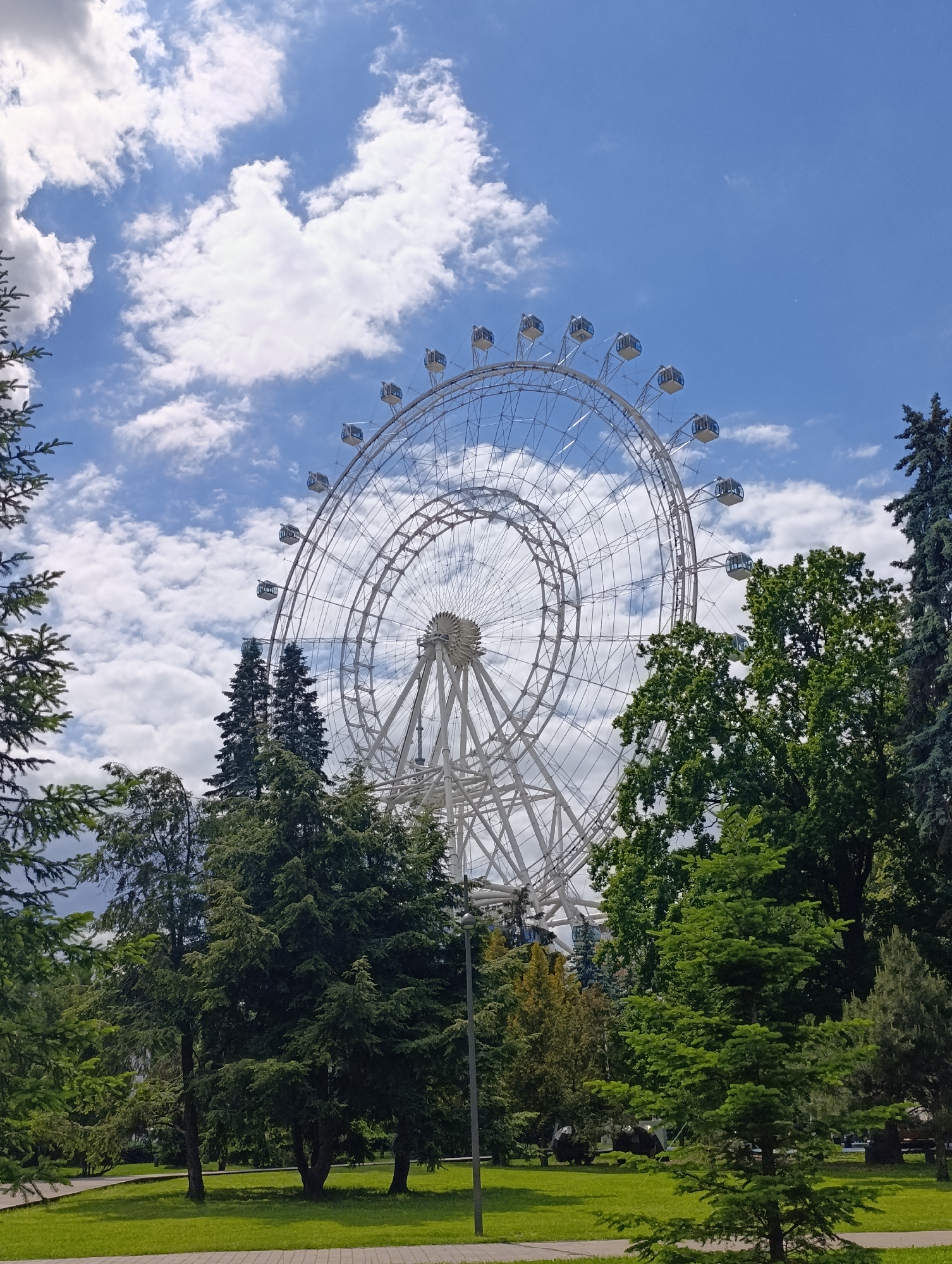 A large, prominent white Ferris wheel dominates the central and upper portions of the image, set against a bright blue sky adorned with scattered white cumulus clouds. The Ferris wheel features a complex skeletal structure of white metal, with multiple circular rings and spokes, and is equipped with numerous enclosed passenger cabins distributed along its outer edge.
The scene is a lush green park or recreational area. Various types of trees, including tall, dark green coniferous trees and lighter green deciduous trees, frame the Ferris wheel from the foreground, sides, and bottom, with their dense foliage partially obscuring the lower parts of the amusement ride. Manicured green grass and paved pathways are visible in the lower section of the image. A dark pole, possibly a light fixture, stands on the left.
The photograph was taken during daytime, under clear and bright conditions, indicative of pleasant weather. The setting suggests a location dedicated to leisure and entertainment, consistent with a park environment. Based on the provided context, the location is Moscow, Russia, implying this could be a known landmark like the "Sun of Moscow" Ferris wheel. There are no visible people or discernible text in the image.