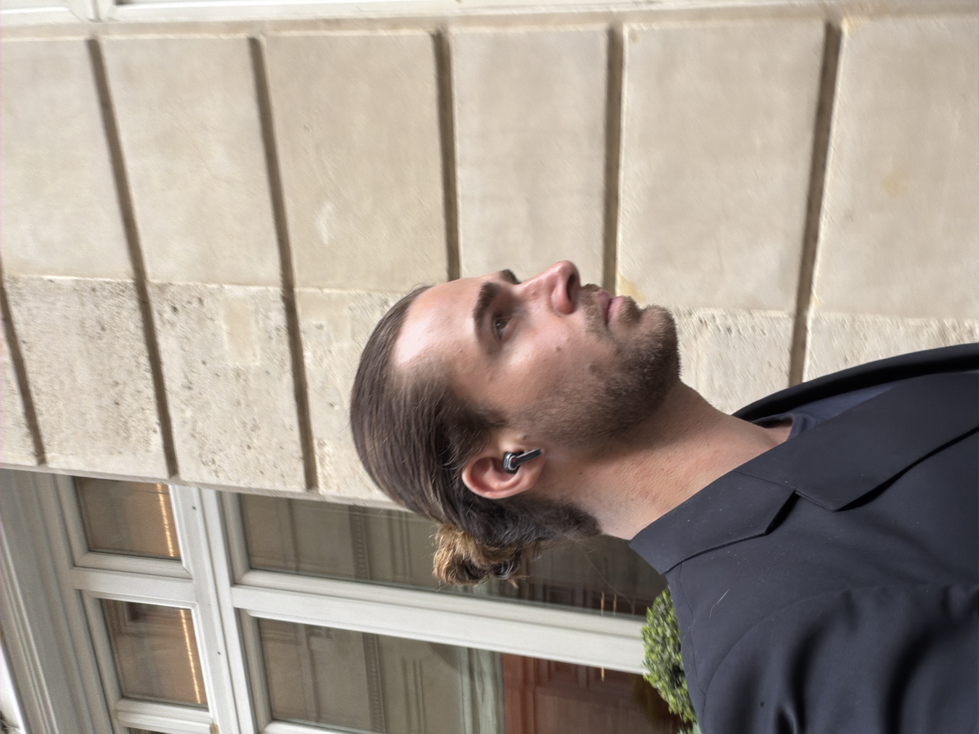 The image features a man in profile, looking upwards with a neutral expression. He has long, dark hair tied back in a small bun at the nape of his neck, and a short beard or stubble. A distinct, dark-colored earbud is visible in his right ear. He is dressed in a dark suit jacket over what appears to be a dark undershirt.
The setting is outdoors, next to a building. The background consists of a light-colored, possibly stone or stucco, building facade with prominent architectural paneling, typical of Haussmannian architecture found in Paris, France. Below the man's head, part of a white-framed window with multiple panes is visible. Subtle green foliage or shrubbery can be seen in the lower right corner, suggesting urban landscaping.
The activity depicted is the man gazing upwards, possibly observing something high on the building or in the sky. The presence of the earbud suggests he might be listening to audio or on a call. The scene is illuminated by bright daylight, indicated by the strong shadows cast by the building's architectural details, implying clear and sunny weather. There is no visible text in the image.