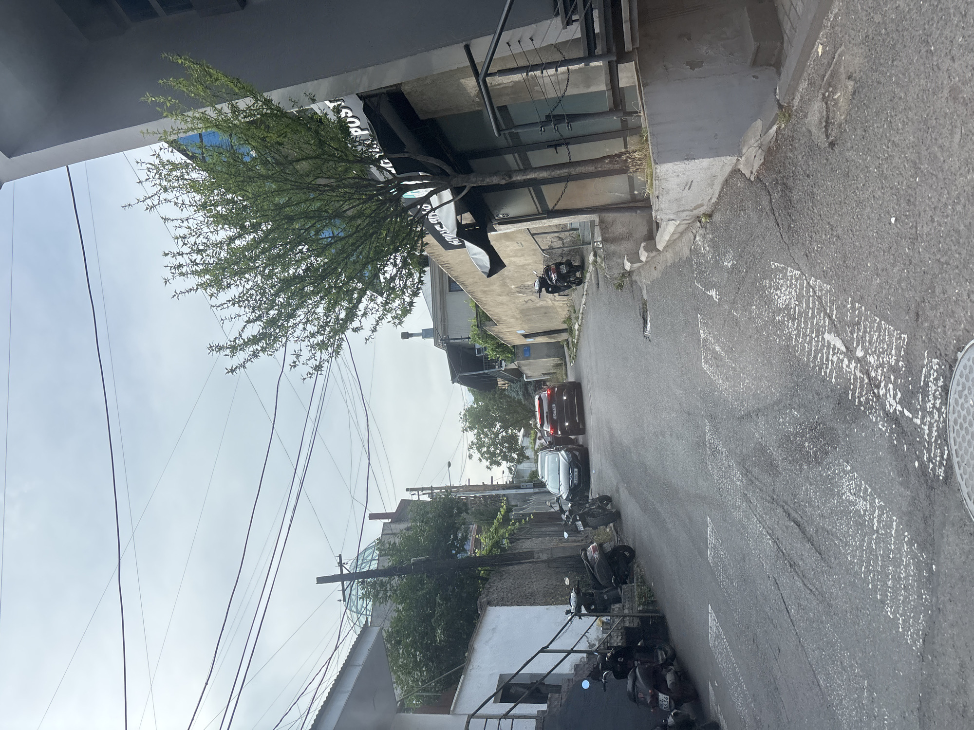 The image depicts a street scene, likely in Tbilisi, Georgia, with a focus on urban infrastructure and elements of daily life. The perspective is from above, looking down an asphalt road lined with buildings and parked motorcycles. The weather appears overcast, suggesting a cloudy day.
Prominent objects in the scene include several parked motorcycles of various colors and models, indicating a common mode of transportation in the area. A dark-colored car is driving down the street, with another car stopped behind it. The buildings on either side of the street vary in architectural style, with some appearing to be older, concrete structures, while others have more modern elements like large glass windows. A prominent tree with lush green foliage grows near one of the buildings, its branches reaching towards the sky. Overhead, a complex network of power lines crisscrosses the frame, a common sight in urban environments.
There are no people clearly visible in the image, but the presence of parked vehicles and the general streetscape suggest an inhabited area. One building has a sign with partially visible text that appears to read "POST." The overall impression is one of an ordinary day in a city, with everyday objects and infrastructure occupying the frame. The lighting and sky suggest it is daytime, but not necessarily bright sunlight.