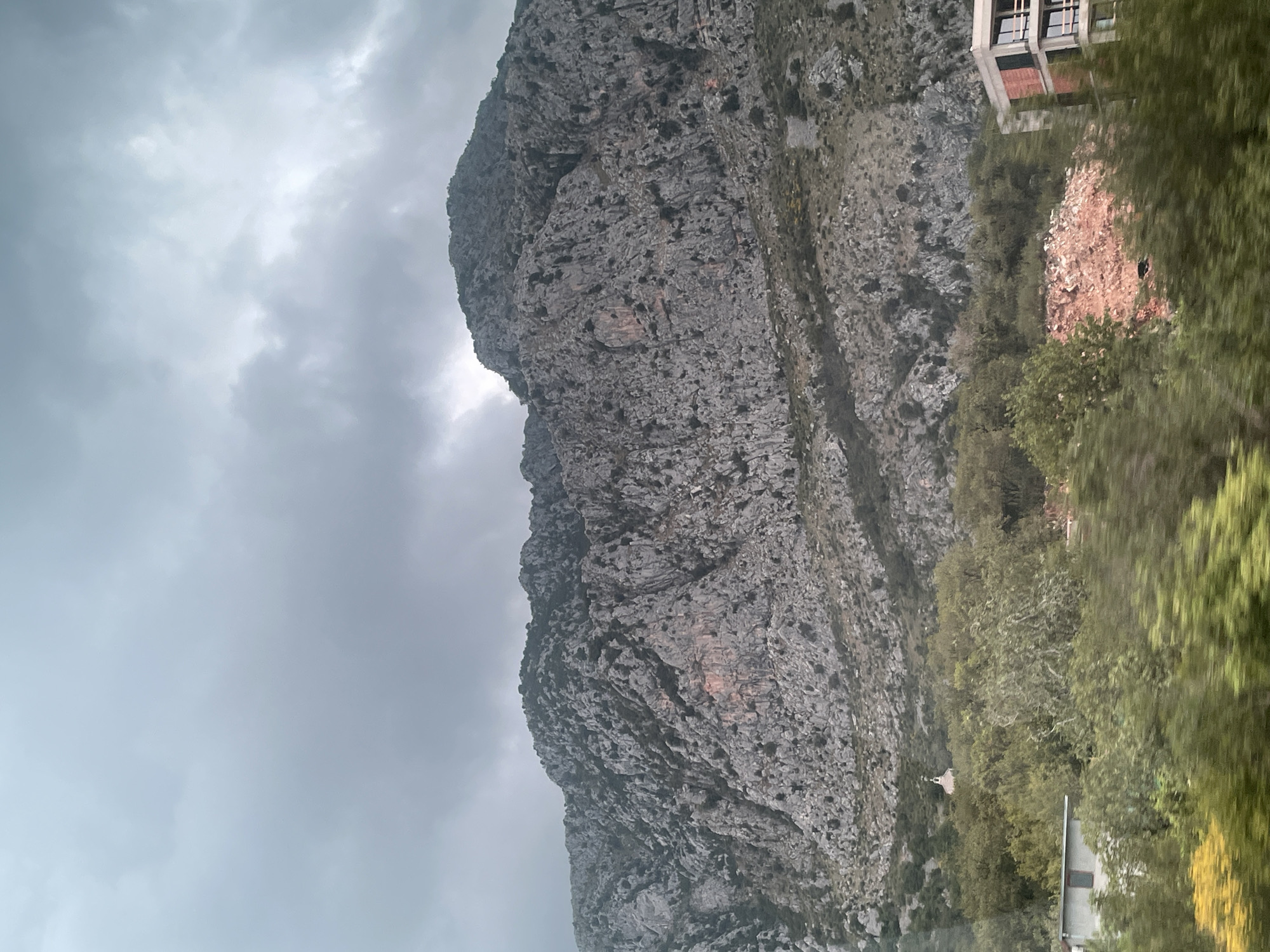 The image displays a vast, rugged mountain range under an overcast sky. The mountain, composed of dark grey, craggy rock, dominates the left and central parts of the frame, exhibiting a textured surface with numerous crevices and scattered patches of dark vegetation. The sky is uniformly cloudy and light grey, suggesting an overcast day with diffused lighting.
On the right side of the image, dense green foliage and trees are prominent in the foreground and midground, with some blur indicating potential motion at the time of capture. Towards the upper right, a modern or unfinished building, likely concrete with visible window frames, is partially visible. Below this structure, a mound of reddish-brown debris or earth is present. Further down on the right, nestled amongst the trees at the base of the mountain, a small, white, almost chapel-like structure with a conical roof can be discerned. There are no people visible, nor is any text present. The scene captures a natural landscape with sparse human structures, possibly in a mountainous Mediterranean or similar region during an overcast day.