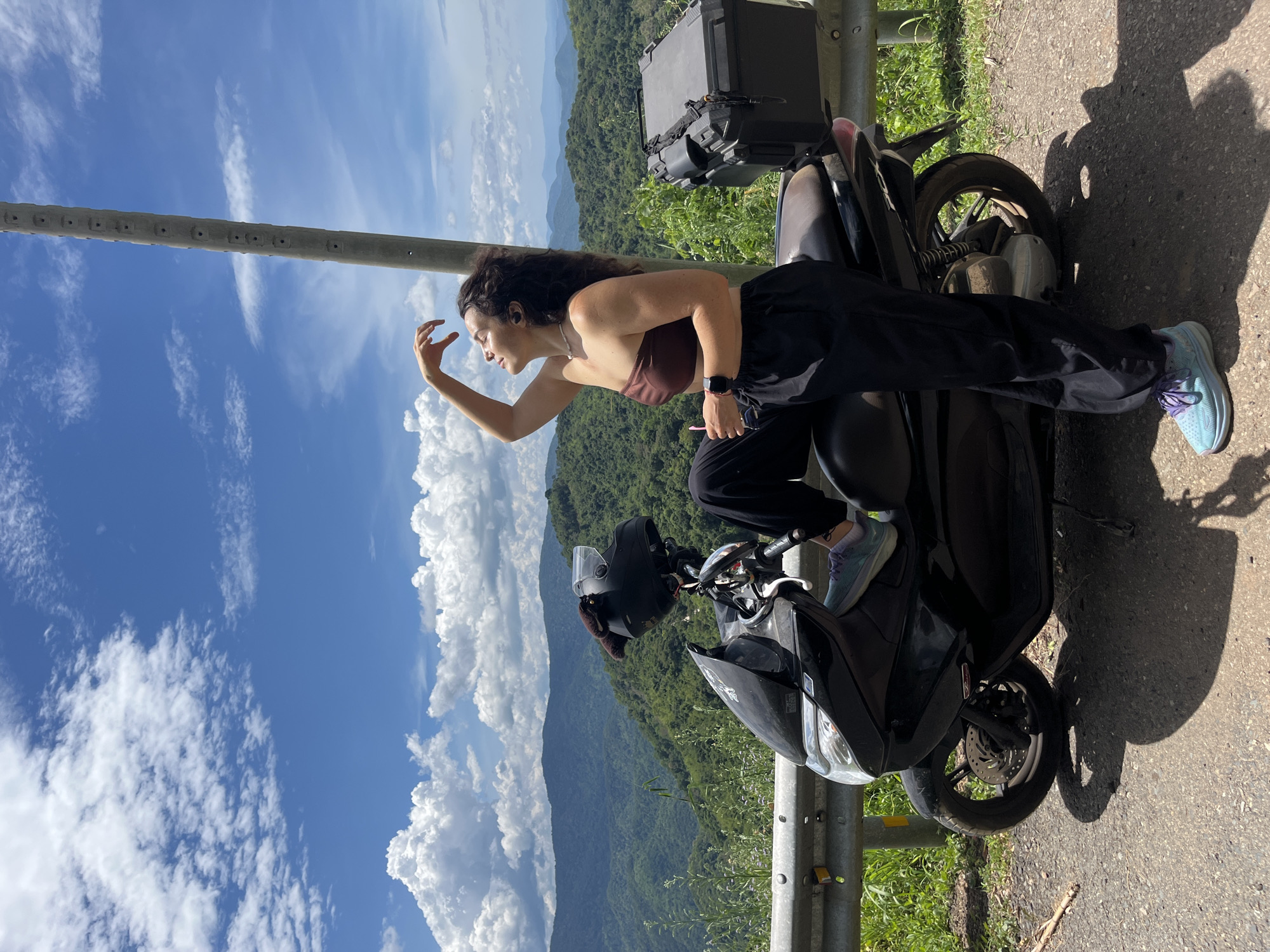 A young woman is posing on a black scooter parked on a road in Son Dien, Vietnam. She is wearing a brown bandeau top, black pants, and blue sneakers with purple laces. Her right leg is propped up on the scooter, and she is holding a pink straw in her hand, touching her forehead. The scooter has a black helmet strapped to the back and a black storage box. The background features a scenic view of lush green mountains under a blue sky with white clouds. A concrete utility pole stands to the right of the woman.