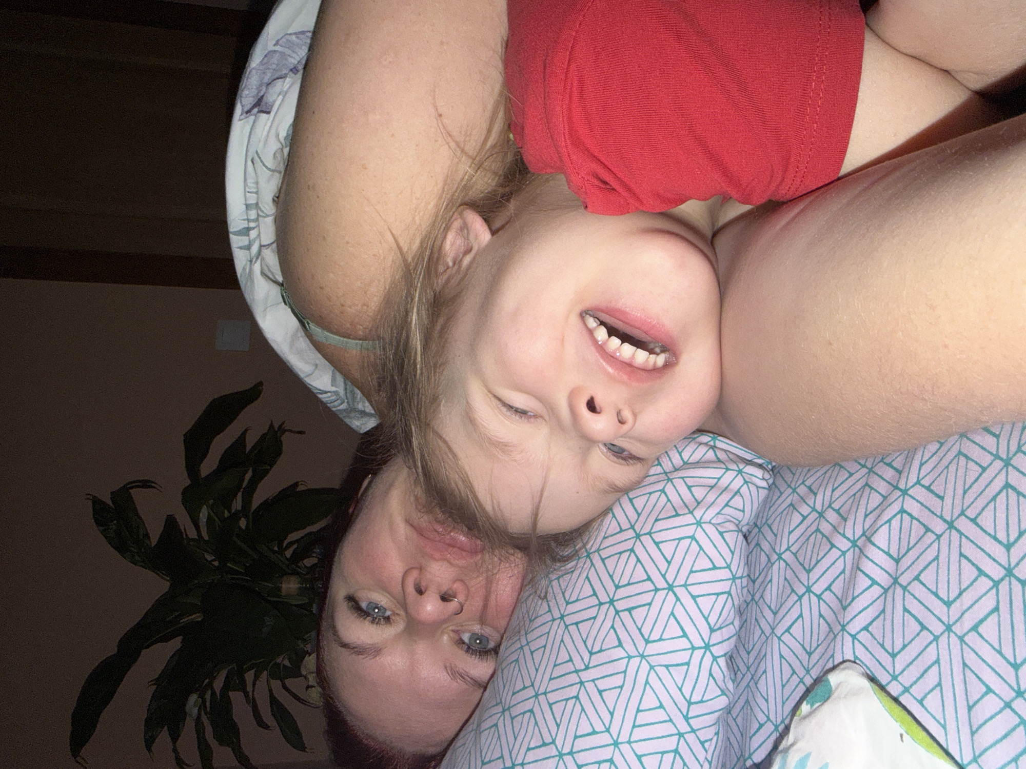 A mother and child are pictured in an indoor setting, likely in Chaikovsky, Russia. The mother is lying down, with a pillow featuring a light blue and white geometric pattern beneath her head. A child in a red shirt is positioned above her, possibly lying on her side. Both are smiling. To the left of the mother's head is a potted plant with green leaves. The background is a light brown wall.