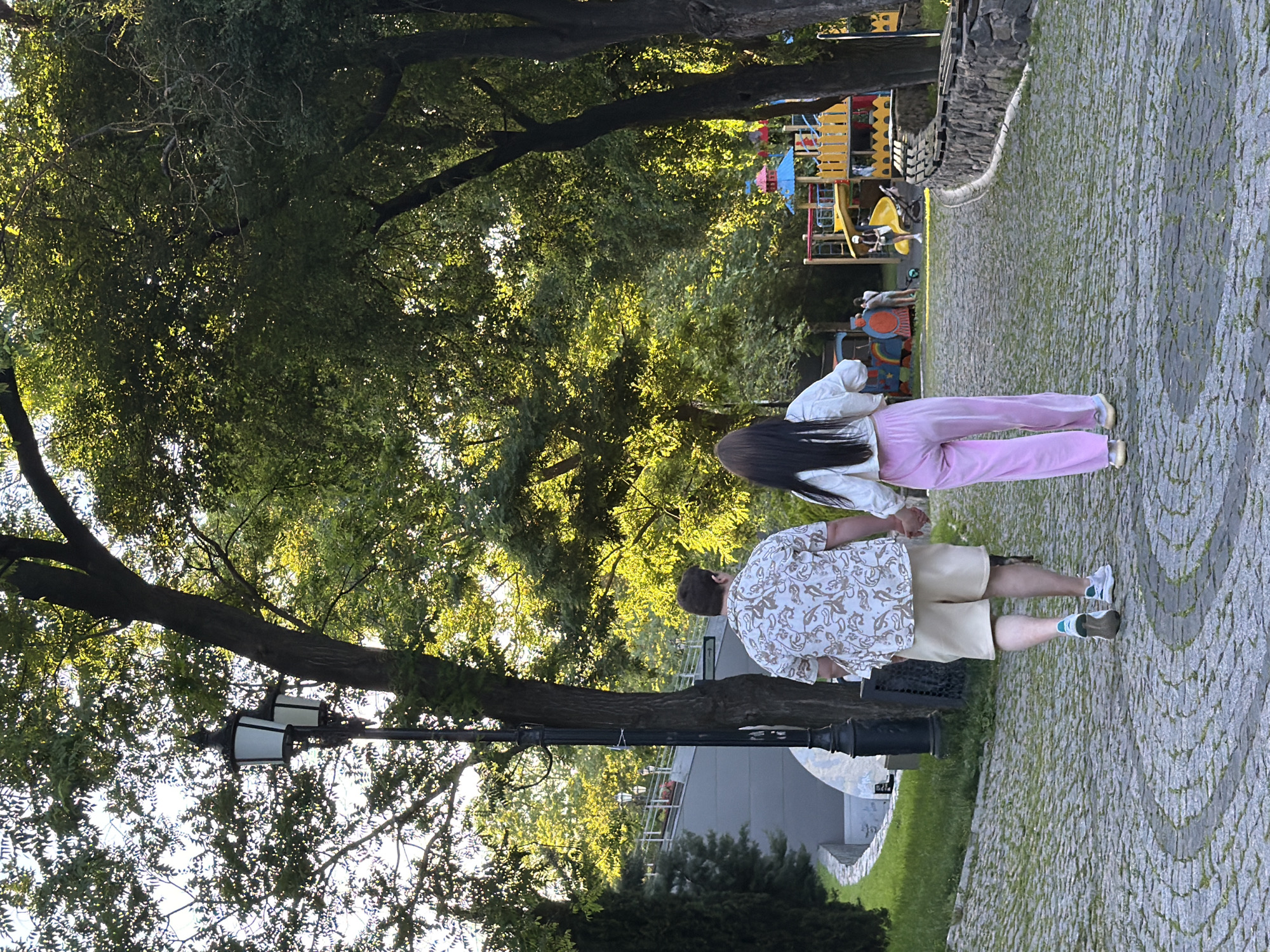 A couple walks hand-in-hand down a cobblestone path in a park in Unknown, Unknown. The man is wearing a light-colored patterned short-sleeved shirt and beige shorts, while the woman is wearing a white long-sleeved shirt and pink pants. They are walking away from the camera, towards a playground visible in the background. The park is lush with green trees, and a vintage-style lamp post stands to the left of the path. In the distance, there is a curved pedestrian bridge. The lighting suggests it is late afternoon.