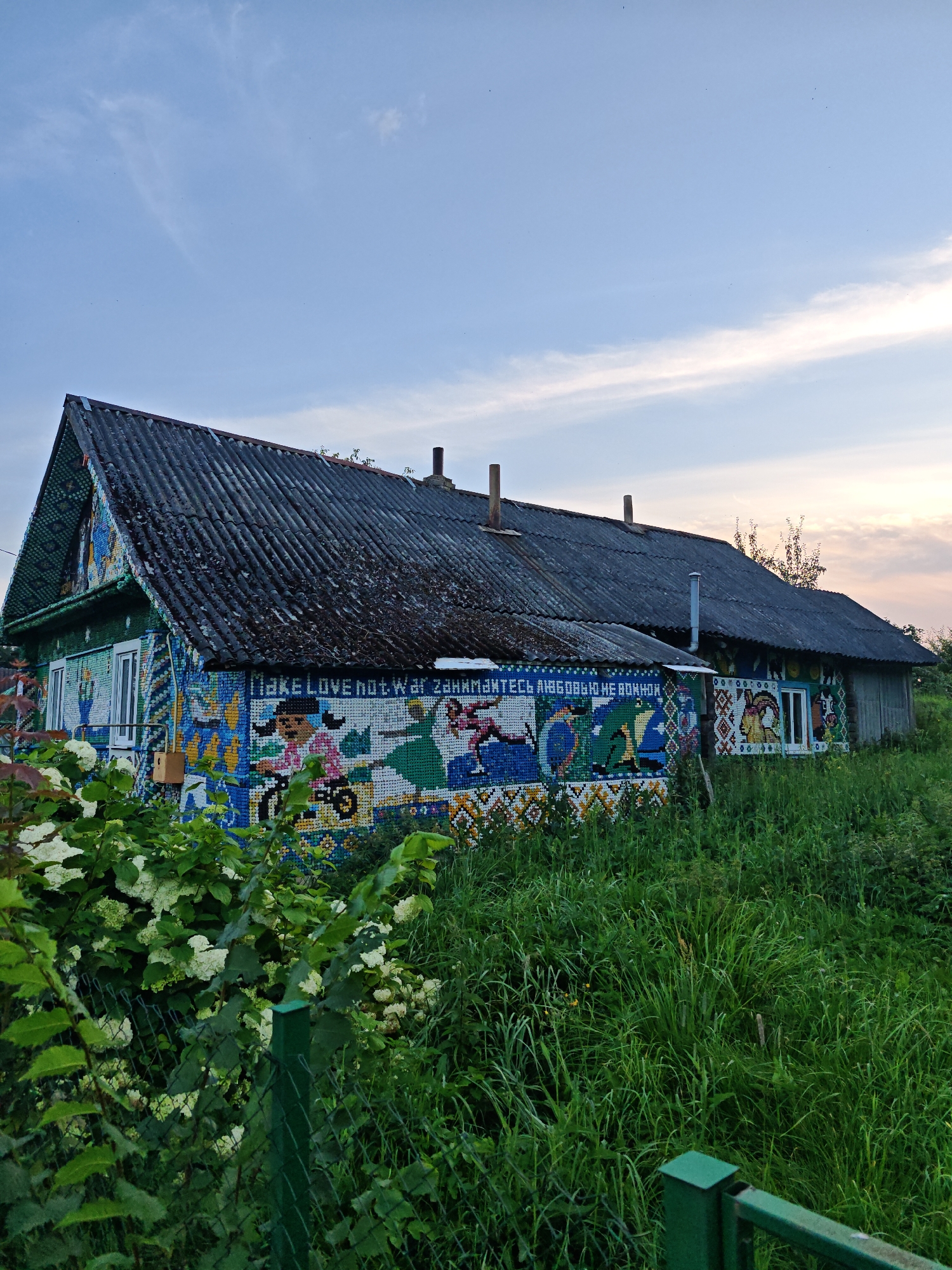 The image shows a house decorated with a mosaic of bottle caps, located in the Валожынскі сельскі Савет, Belarus. The house has a dark, corrugated roof and is covered in colorful bottle cap art on its walls. The art depicts various images and text, including what appears to be "Make Love not War" in English and Cyrillic script. The house is surrounded by lush green grass and foliage, and a fence is visible in the foreground. The sky is a pale blue with some wispy clouds, suggesting it is either early morning or late evening.