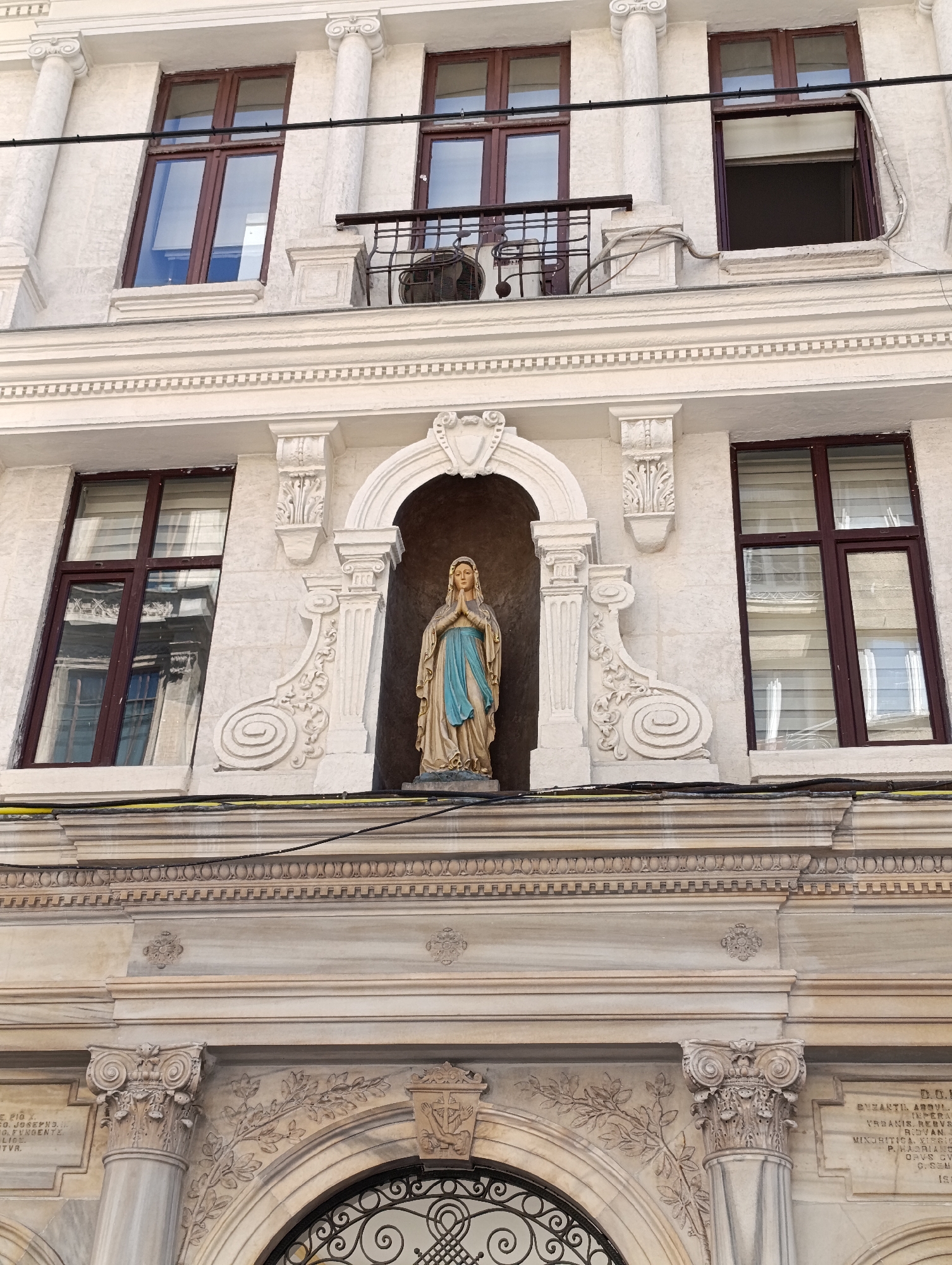The image shows a close-up, low-angle shot of a building facade in Beyoğlu, Turkey. The facade is predominantly white with decorative architectural elements. In the center, a statue of the Virgin Mary, dressed in a blue and beige robe, stands in a niche above a carved ornamental molding. She is depicted with clasped hands and a serene expression. Flanking the niche are ornate white stucco decorations, including volutes and floral motifs. Above the niche, there are two large windows with dark brown frames, reflecting the sky and surrounding buildings. To the left and right of the niche, there are additional windows with similar dark brown frames. A black electrical wire runs horizontally across the upper part of the facade, just below the top row of windows. A metal balcony railing is visible on the right side, with an air conditioning unit mounted below it. The lower portion of the image reveals more of the building's intricate stonework, including columns with Corinthian capitals and carved friezes. An arched entryway with decorative ironwork is visible at the very bottom.