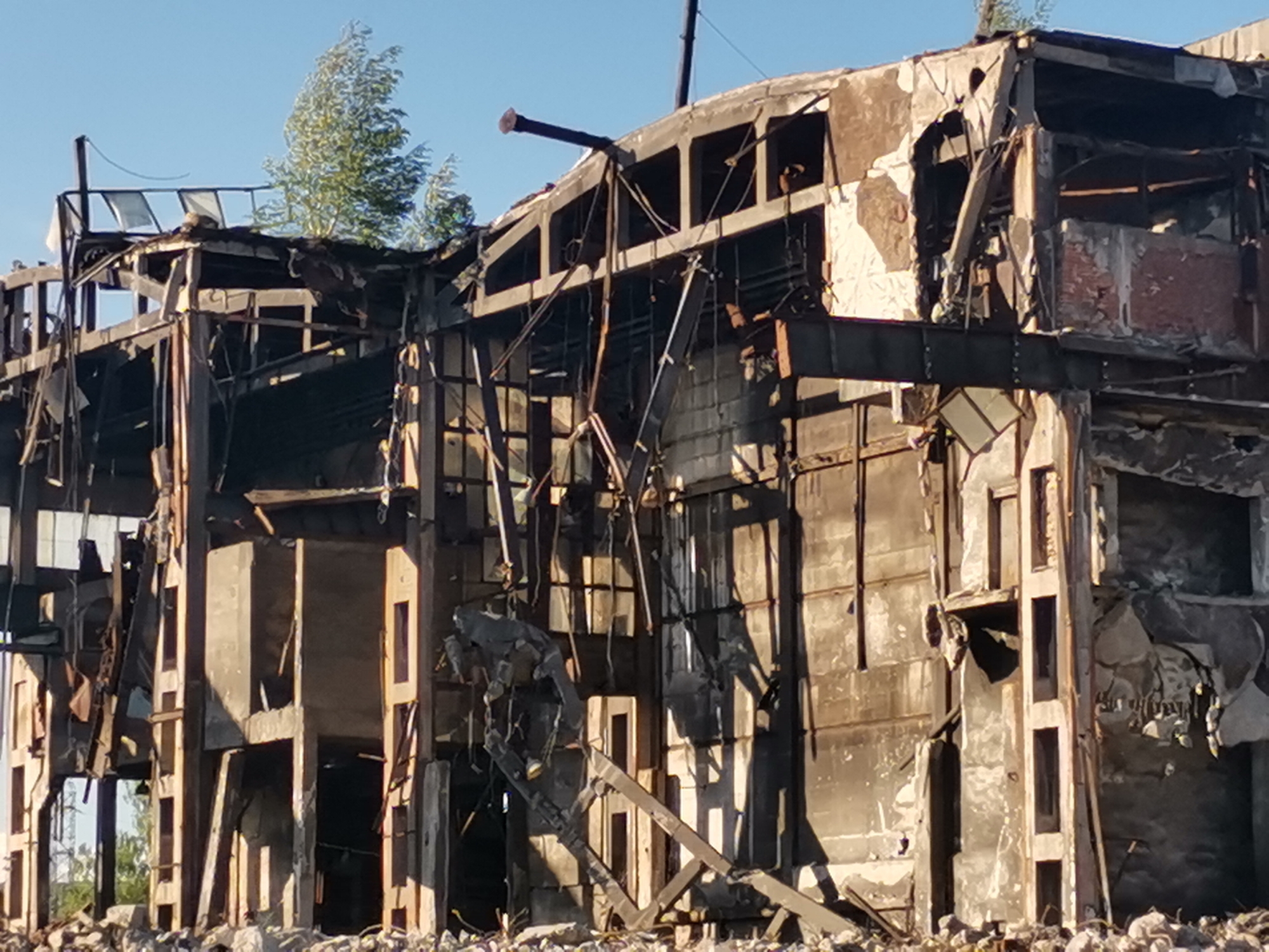 The image depicts the skeletal remains of a severely damaged building in Zhivkovo, Bulgaria. The structure appears to be a multi-story building constructed primarily of concrete and steel. The facade is largely destroyed, revealing interior spaces and structural elements. Charred materials indicate extensive fire damage. The surrounding ground is covered with rubble and debris from the building's collapse. Some vegetation is visible growing within and around the ruins. No people are visible in the frame.
