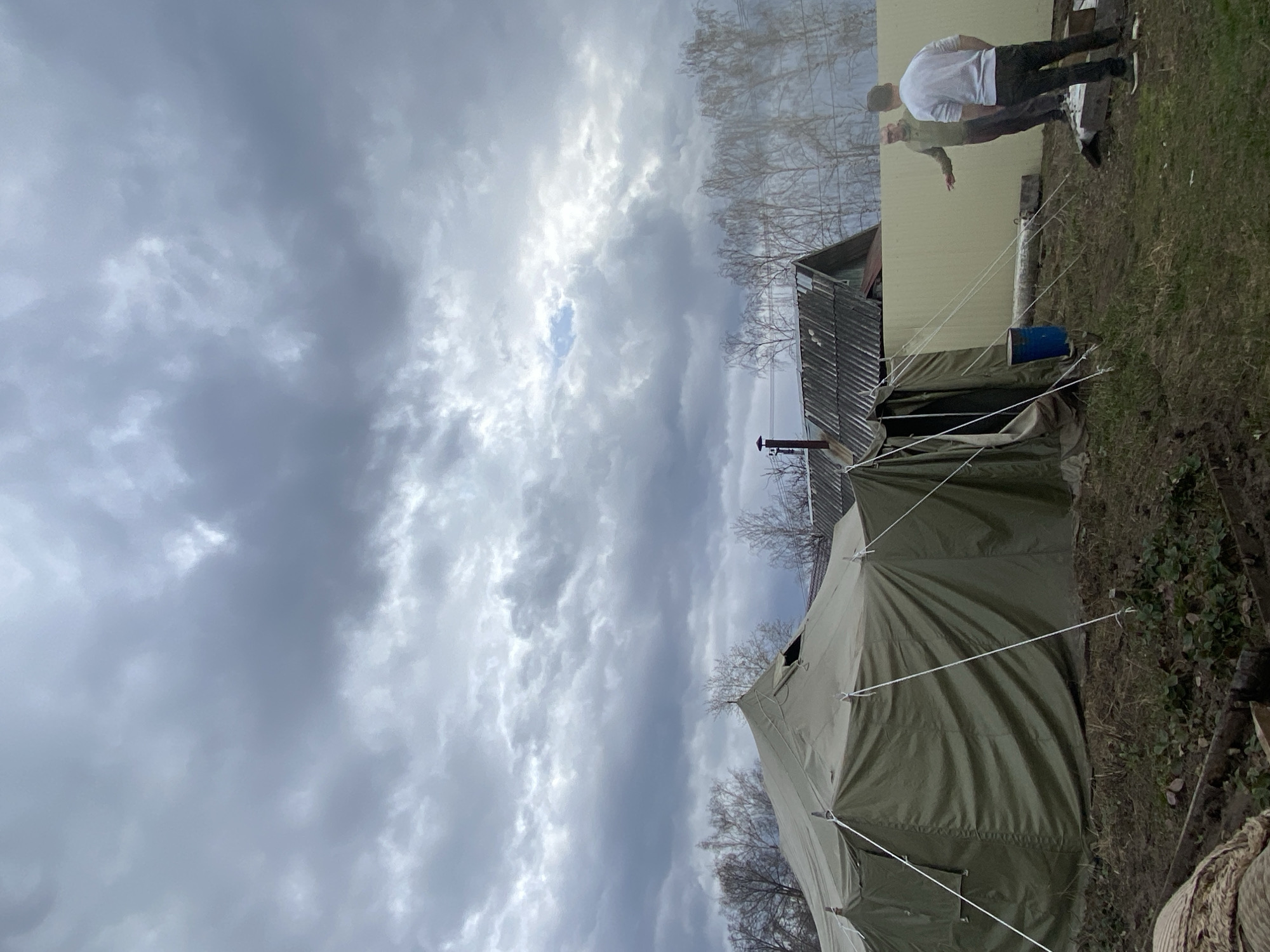 The image shows a campsite under a cloudy sky. A large, green tent dominates the foreground, with several white guy-lines anchoring it to the ground. Behind the tent is a weathered, gray-roofed building, possibly a shed or small house. Two men are standing near a yellow building with one in a white shirt and the other in a dark colored jacket or shirt. The location is suggested to be Chaikovsky, Russia. The ground appears to be a mix of dirt and grass. A blue barrel is placed on the ground near the shed. Bare tree branches are visible in the background, indicating it may be late autumn or early spring.