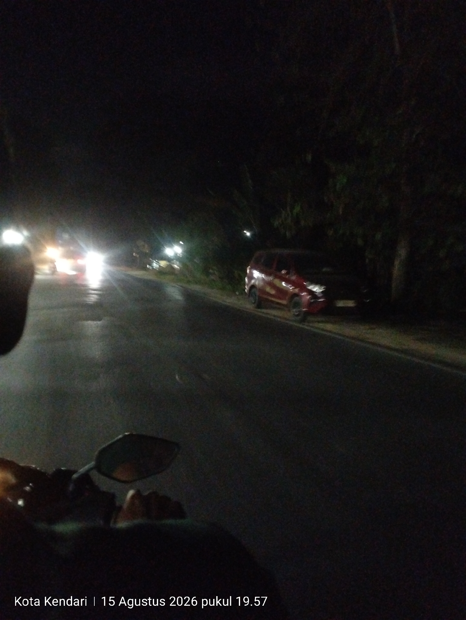 The image depicts a nighttime scene on a road in Kendari, Indonesia, as indicated by the text overlay "Kota Kendari | 15 Agustus 2026 pukul 19.57". The perspective appears to be from a moving vehicle, likely a motorcycle, with a rearview mirror visible in the foreground. The road surface is wet, reflecting the lights of passing vehicles. In the distance, several cars with illuminated headlights and taillights are visible, suggesting traffic. On the right side of the road, a red minivan is parked, partially obscured by darkness and trees. The environment is characterized by a dark, tree-lined road, typical of an urban or suburban setting at night.