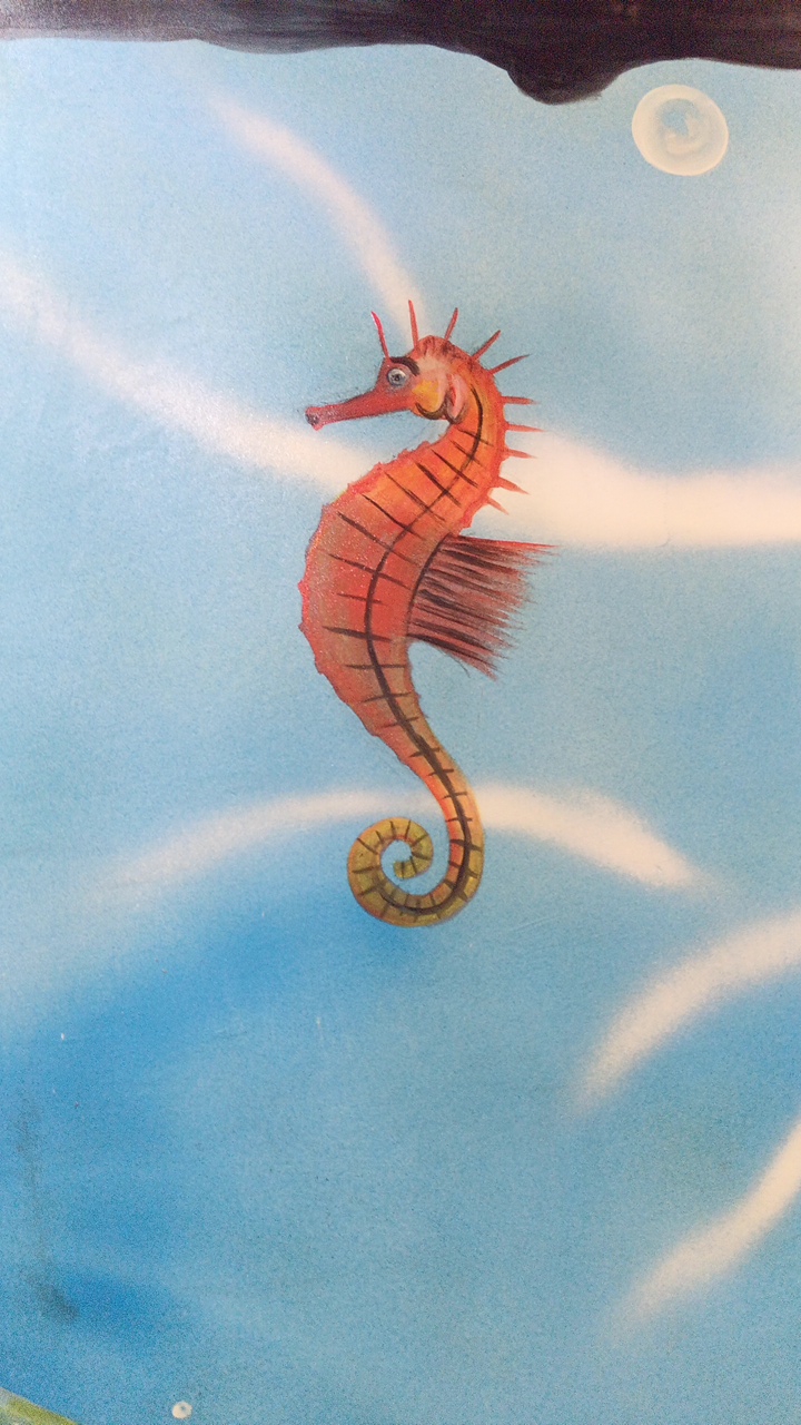 A painted mural depicts a vibrant orange seahorse with black stripes swimming in a bright blue aquatic environment. The seahorse is curled into a spiral shape, with its head facing left. Wispy white clouds or currents surround the seahorse. At the top of the mural, a dark, amorphous shape suggests an underwater cave or overhang. A pale, circular object, possibly a stylized moon or bubble, is visible in the upper right portion of the image. The overall style is illustrative and fantastical. The image is a photograph of a mural and does not show any identifiable elements of the city of Balykchy, Kyrgyzstan.