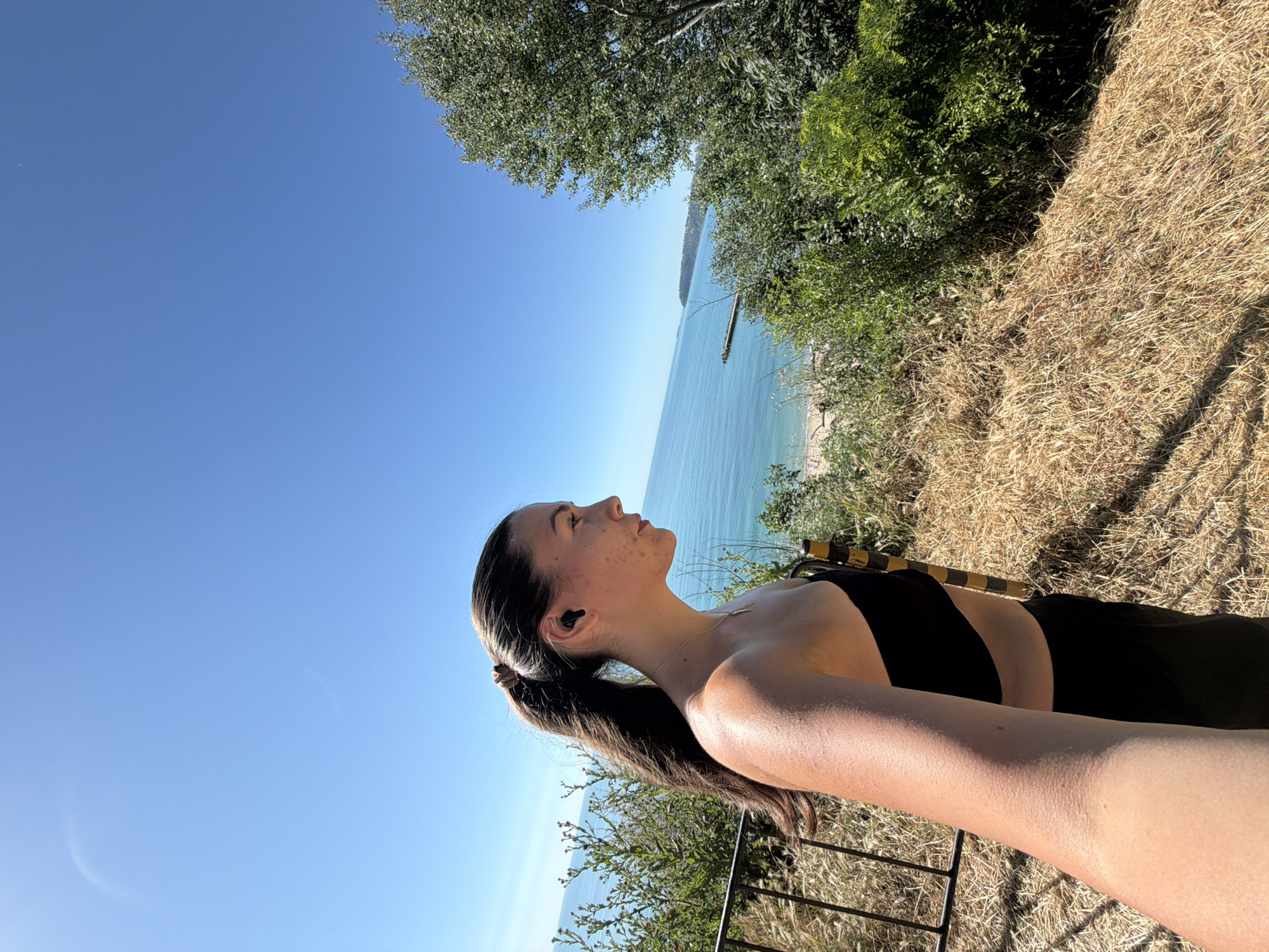 A young woman with dark hair in a ponytail, wearing a black strapless top and black pants, stands on a hill overlooking the sea. She has an earpiece in her right ear and appears to be looking out at the water. The area around her is covered in dry grass and some green bushes. In the background, the blue sea stretches to the horizon, with a distant landmass and a pier. The sky is clear and blue. The scene is set on a sunny day. This viewpoint is likely from a location in Varna, Bulgaria, such as the Sea Garden or a coastal path offering panoramic views of the Black Sea.