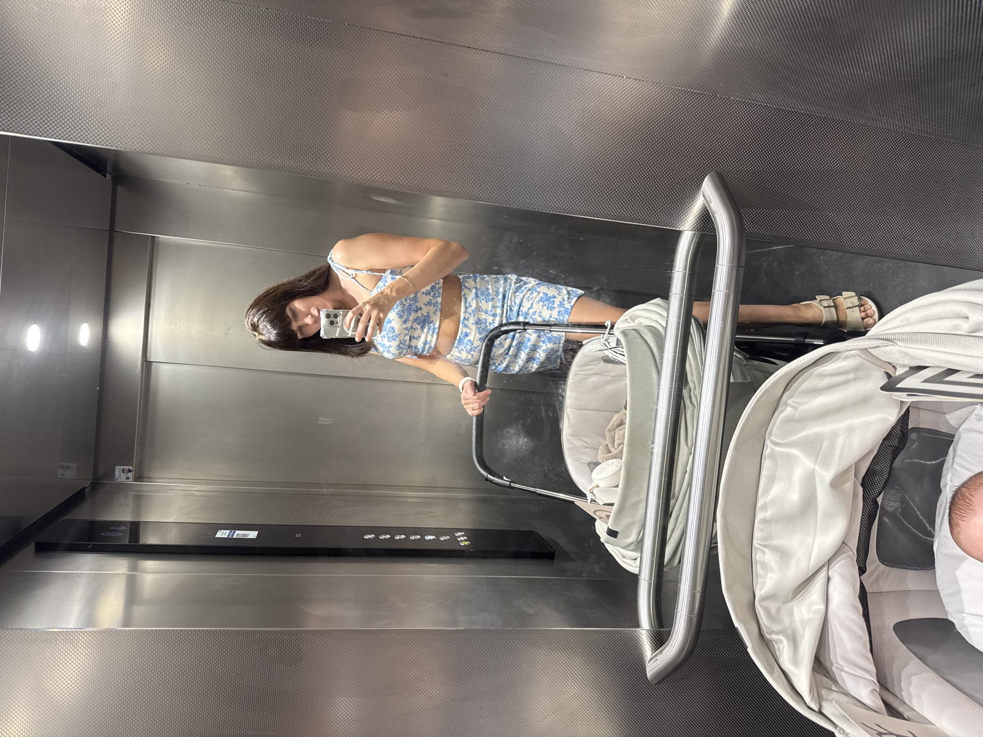 The image shows a woman taking a mirror selfie inside an elevator. She is wearing a blue and white floral print crop top and skirt. She is holding a smartphone in her right hand and her left hand is resting on the handle of a stroller. A baby is visible in the stroller. The elevator has stainless steel walls and a black panel with elevator buttons. The floor appears to be dark. The image does not contain any visual elements that would indicate the location of Isola delle Femmine, Italy.