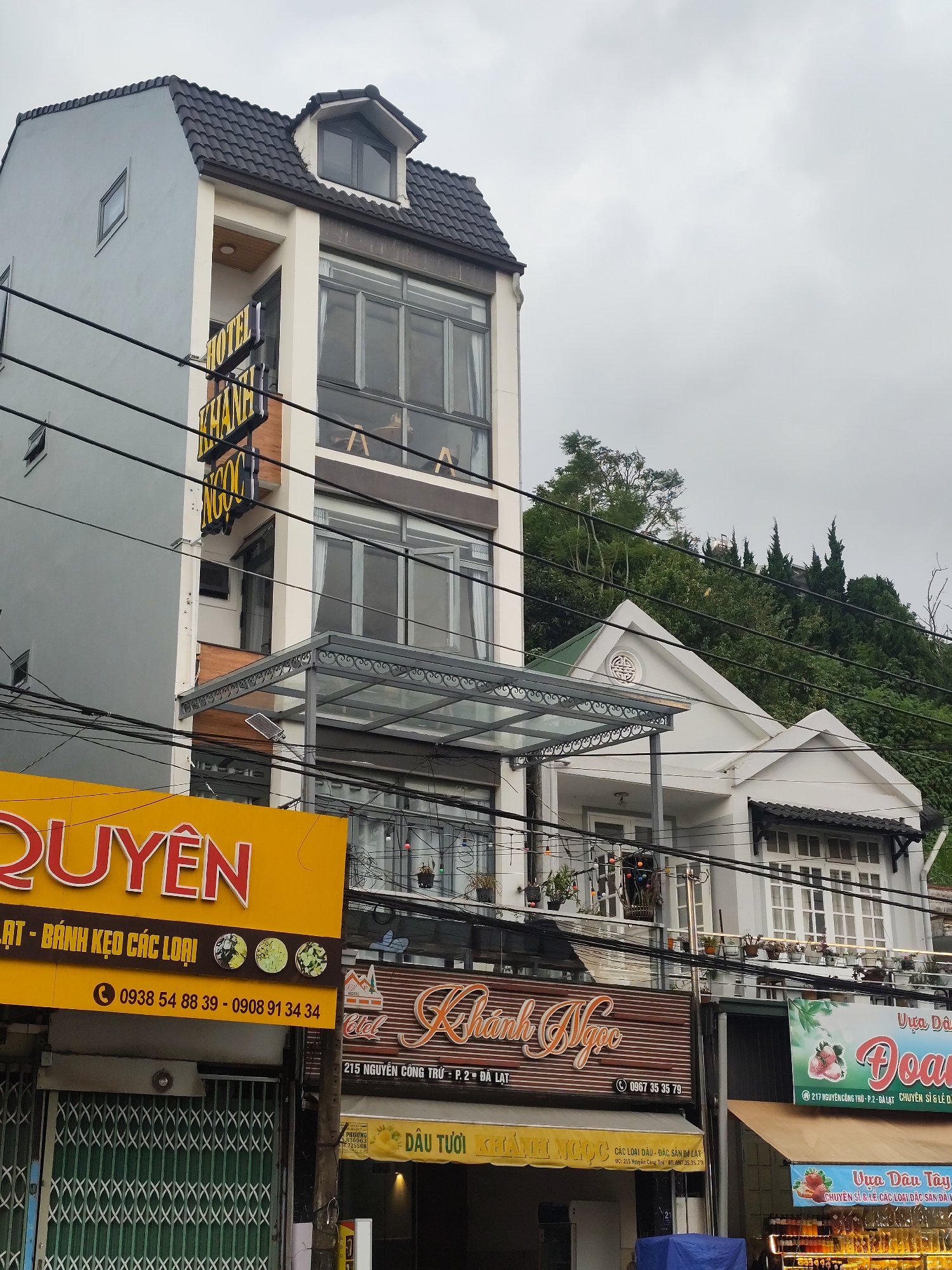 This media file captures a street view in Da Lat, Vietnam, on a cloudy day. The foreground is dominated by storefronts and signage. A bright yellow sign with red lettering that reads "QUYÊN" is prominent, advertising "LẠT - BÁNH KẸO CÁC LOẠI" (presumably types of candy and pastries) and providing phone numbers. To the right, a horizontal sign in cursive gold lettering on a brown background says "KHÁNH NGỌC" with a phone number and the address "215 NGUYỄN CÔNG TRỨ - P. 2 - ĐÀ LẠT". Further to the right, a green and white sign displays "Vựa Dâu" and "Doa" with images of strawberries, indicating a strawberry vendor.
Behind these signs, a multi-story building with a modern facade, featuring large glass windows and a dark tiled roof, stands tall. The building appears to be a hotel, with a sign partially visible that reads "HOTEL KHANH NGỌC". A smaller white building with a pitched roof and an ornamental circular detail is situated to the right of the hotel, partially obscured by power lines.
The background is a densely forested hillside, typical of Da Lat's topography. The sky is overcast, suggesting a subdued or potentially rainy atmosphere. The scene depicts a commercial street, likely a bustling area for local produce and goods, with an emphasis on "ĐẶC SẢN ĐÀ LẠT" (Da Lat specialties). The power lines crisscrossing the frame are a common urban element in the scene. No people are clearly visible in the frame.