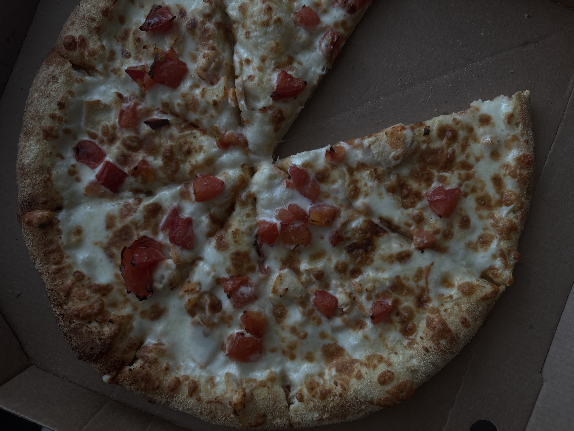 An overhead shot displays a partially eaten pizza resting inside an open, plain brown cardboard box. The pizza, which appears to be freshly baked, takes up most of the frame. One slice is visibly missing, creating a triangular void in the otherwise circular arrangement of the remaining slices.
The pizza is generously topped with melted, creamy white cheese, likely mozzarella, covering the entire surface up to the golden-brown crust. Scattered across the cheese are numerous diced red tomatoes, some of which show slight browning or charring on their edges from baking. The crust is thick and appears to have a slightly bready texture, with a visible dusting of flour or cornmeal on its surface, particularly along the rim.
The lighting in the image is dim and soft, casting subtle shadows and giving the scene a cozy, possibly evening atmosphere. The overall color palette is warm, dominated by the browns of the crust and box, the whites and light yellows of the cheese, and the vibrant red of the tomatoes.
No specific activity beyond the presence of the pizza and the missing slice is depicted. There are no people visible. The cardboard box is generic, lacking any discernible logos, branding, or text. There are no visible outdoor elements or specific location cues to tie the image to Zheleznogorsk, Russia, or any other particular place.