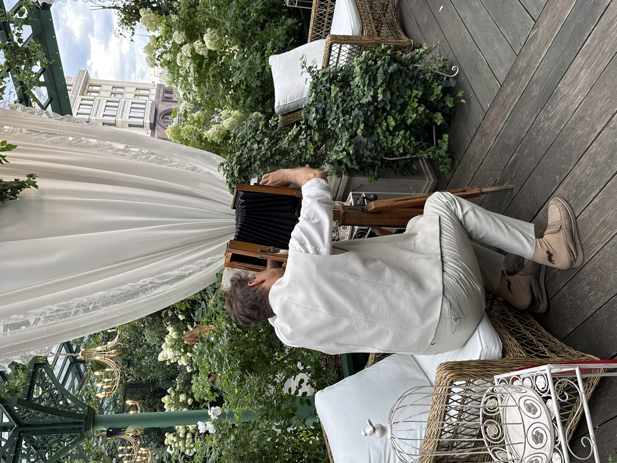 A man is taking a photo with a vintage camera in a garden area. He is wearing a light-colored vest and pants, and brown shoes. The garden has lush green plants and white flowers. There are also wicker chairs and a white birdcage in the scene. In the background, a tall building is visible. The man appears to be focused on his photography, with his hands on the camera. The overall setting suggests an outdoor event or a photographer's studio in a garden. This location is likely within the city of Moscow, Russia, given the architectural style of the building in the background and the commonality of such garden venues in the city.