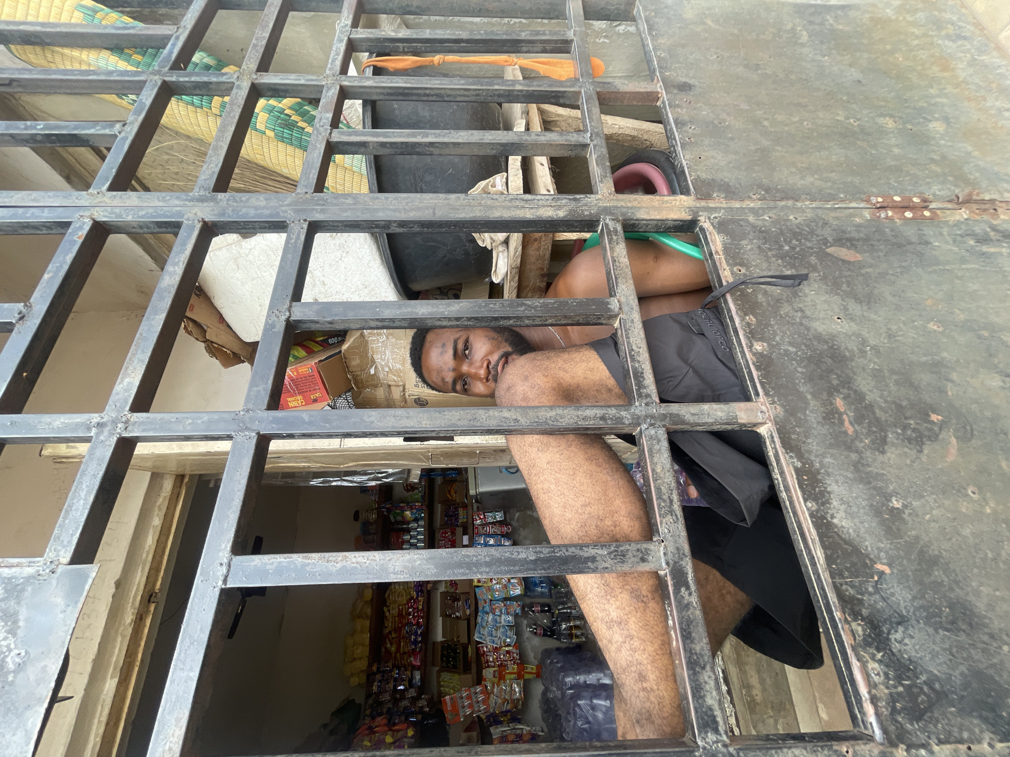 A man is observed looking through a metal grate or window with multiple rectangular openings, which appears to be a security barrier. The metal structure is painted black and shows signs of wear. He is positioned such that his face and part of his upper body are visible.
Through the grate, a shop is visible, filled with various items such as snacks and beverages arranged on shelves. The shop seems to be well-stocked. Miscellaneous items, like woven mats and containers, are visible behind the man. The setting appears to be Lokoja, Nigeria.