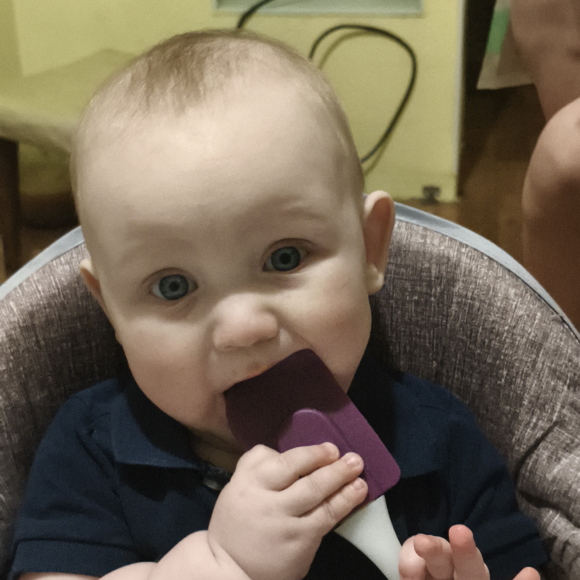 A baby is sitting in a high chair and holding a purple spatula in their mouth. The baby has blue eyes and fair skin. The high chair has a gray fabric seat. The baby is wearing a dark blue shirt. The background is blurry but appears to be a room with light yellow walls.