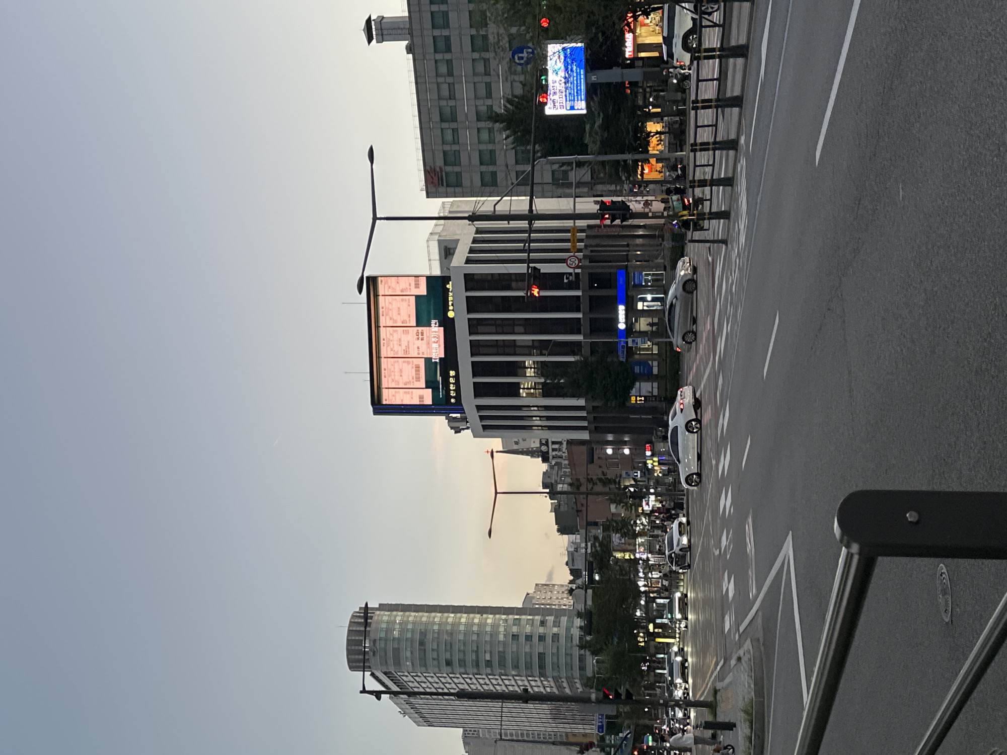 The image captures a street scene in Seoul, South Korea, likely during the evening hours given the sky's twilight hue and the illuminated signage. The foreground shows a street with white lane markings and a pedestrian crossing. Several cars are driving on the road, including a white sedan on the right and a silver sedan on the left. In the midground, a busy intersection is visible with traffic lights, street lamps, and various buildings lining the street. Prominent among these are a tall, cylindrical building with many windows on the left, and a modern building with a large digital billboard at its top, displaying what appears to be advertisements or information. Further buildings, some with illuminated windows and signage, extend into the background. Pedestrians and a few motorcycles can also be observed on the sidewalks and street. The overall impression is of a vibrant urban environment with active traffic and commercial activity.
