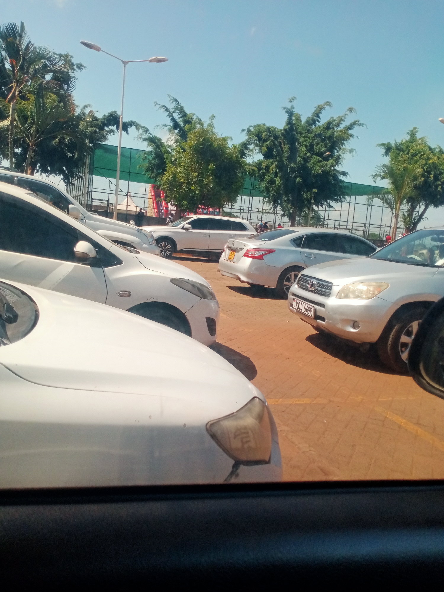 The image shows a parking lot on a sunny day. Several cars are parked close together, including white and silver sedans and an SUV. License plates are visible on some of the vehicles. In the background, trees, a tall streetlight, and a green and white structure (possibly a fence or awning) are visible. The photograph was likely taken from inside a car, as part of a car door is visible in the lower portion of the image. It is unknown if the location is Mugumo-ini ward, Kenya. No people are clearly visible.