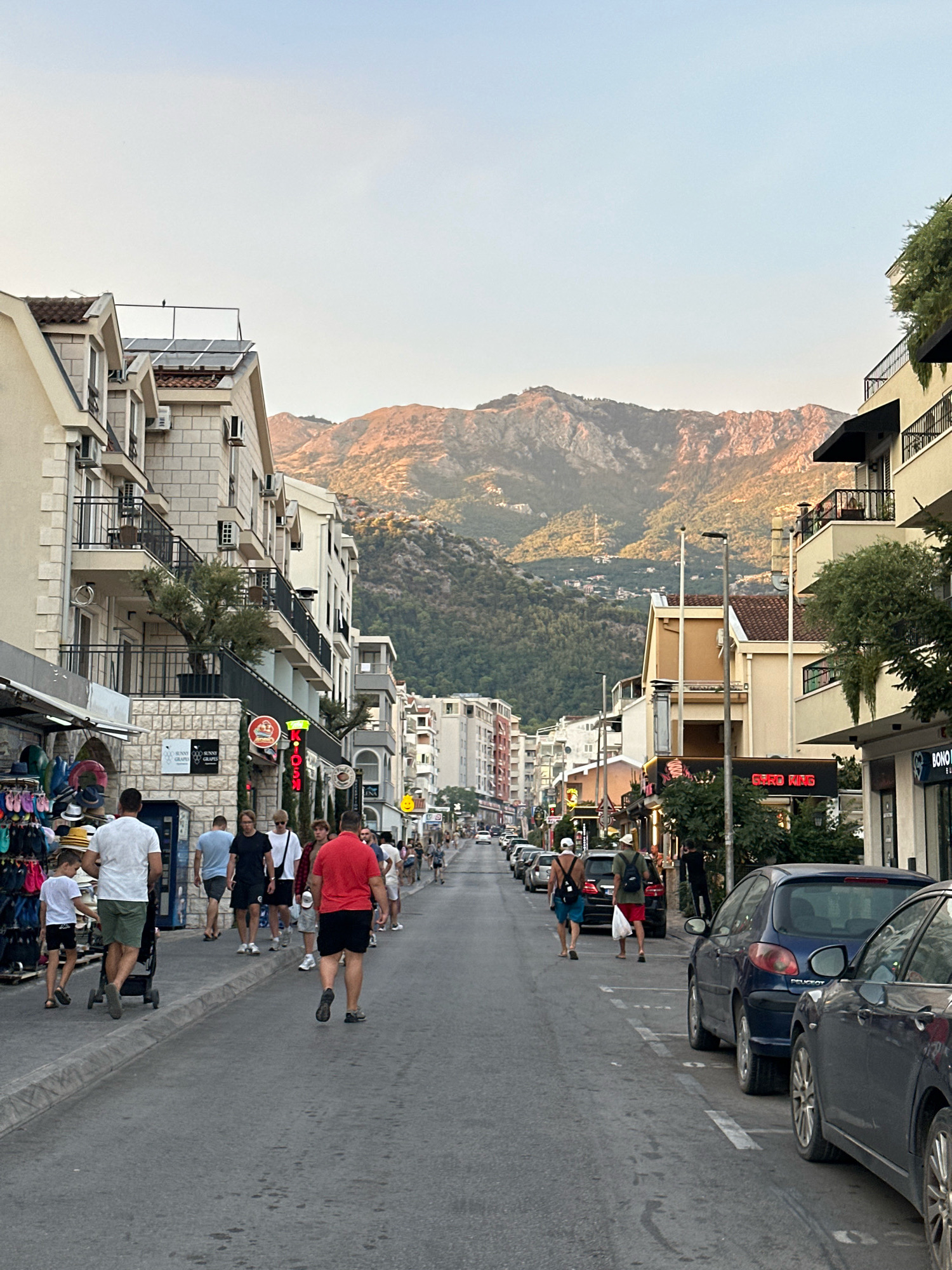 This is a street-level, medium-wide shot taken in Budva, Montenegro. The view is down a busy street lined with multi-story buildings housing shops and residences. The buildings are a mix of architectural styles, with balconies and various window types. Several people are walking along the sidewalk and crossing the street. Cars are parked on both sides of the street and some are driving. In the background, a mountain range is visible, catching the warm light of either sunrise or sunset. The street is paved with asphalt and marked with white lines for parking. Shops on the left side display merchandise like hats and flip-flops. There are streetlights and some trees and greenery. The sky is clear and pale blue.