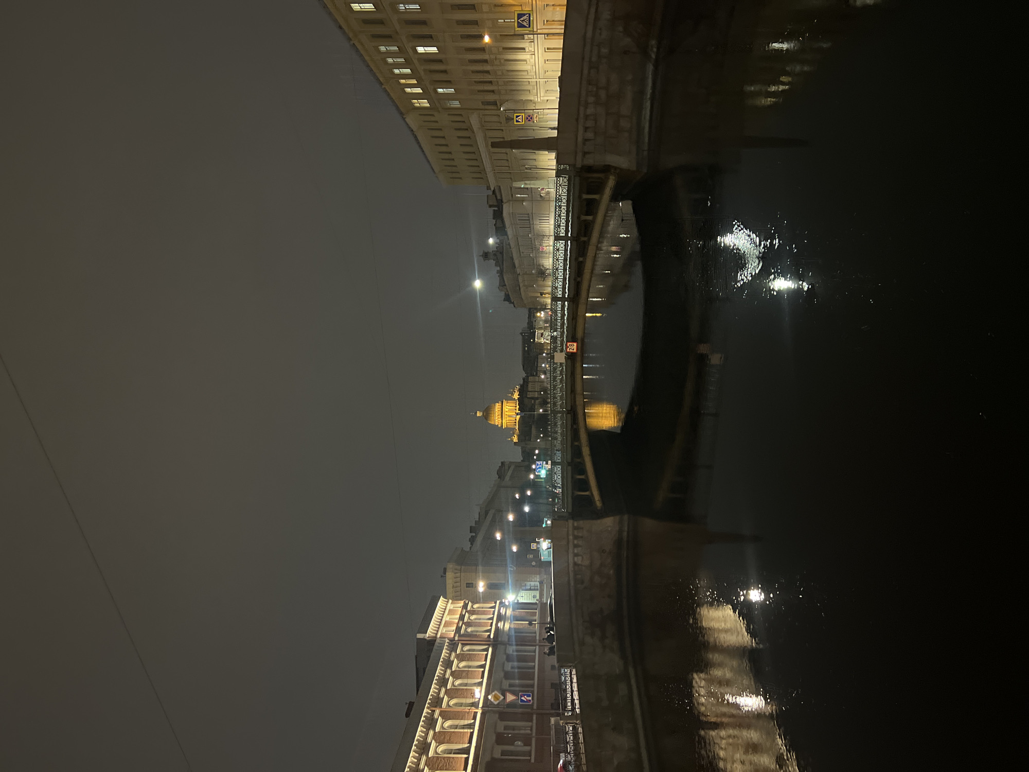 The image displays an urban night scene along the Moika River Embankment in Saint Petersburg, Russia, facing west. Pevchesky Bridge (Singers' Bridge) spans the Moika River in the mid-ground, with the illuminated golden dome of St. Isaac's Cathedral visible in the distant background. The sky is dark and uniformly overcast.
The Moika River, dark and reflecting city lights, occupies the lower portion of the frame. Pevchesky Bridge, a multi-arch structure with intricate dark metal railings, crosses the river, its understructure and arches casting reflections onto the water.
Buildings line both sides of the embankment. On the left, a multi-story building features red-brick upper sections with white architectural trim around arched windows, illuminated by warm yellow light. Several indistinct human figures are visible on this embankment, along with a light-colored vehicle. On the right, another multi-story building with a lighter facade, possibly cream or yellow, and numerous rectangular windows is also illuminated. Numerous streetlights and building lights are active, creating bright points of light and streaks of reflection on the water. Traffic signs, including pedestrian crossing signs, are present on the bridge and embankments.