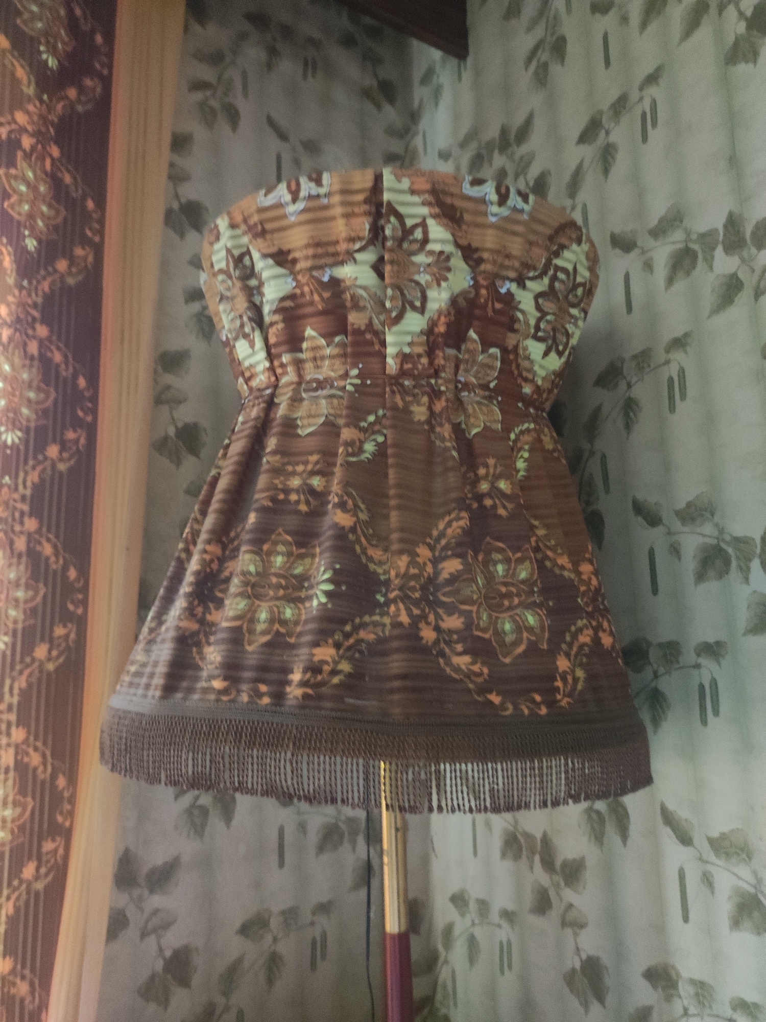 The image shows a close-up of a lampshade with a floral and striped pattern in shades of brown, orange, and green. The lampshade has fringe along its bottom edge. The lampshade is on a lamp with a metallic pole. In the background, there is a curtain with a light green leaf pattern. The lighting suggests an indoor setting. The image does not provide any information about the city of Velikiye Luki, Russia, or any specific location within it.