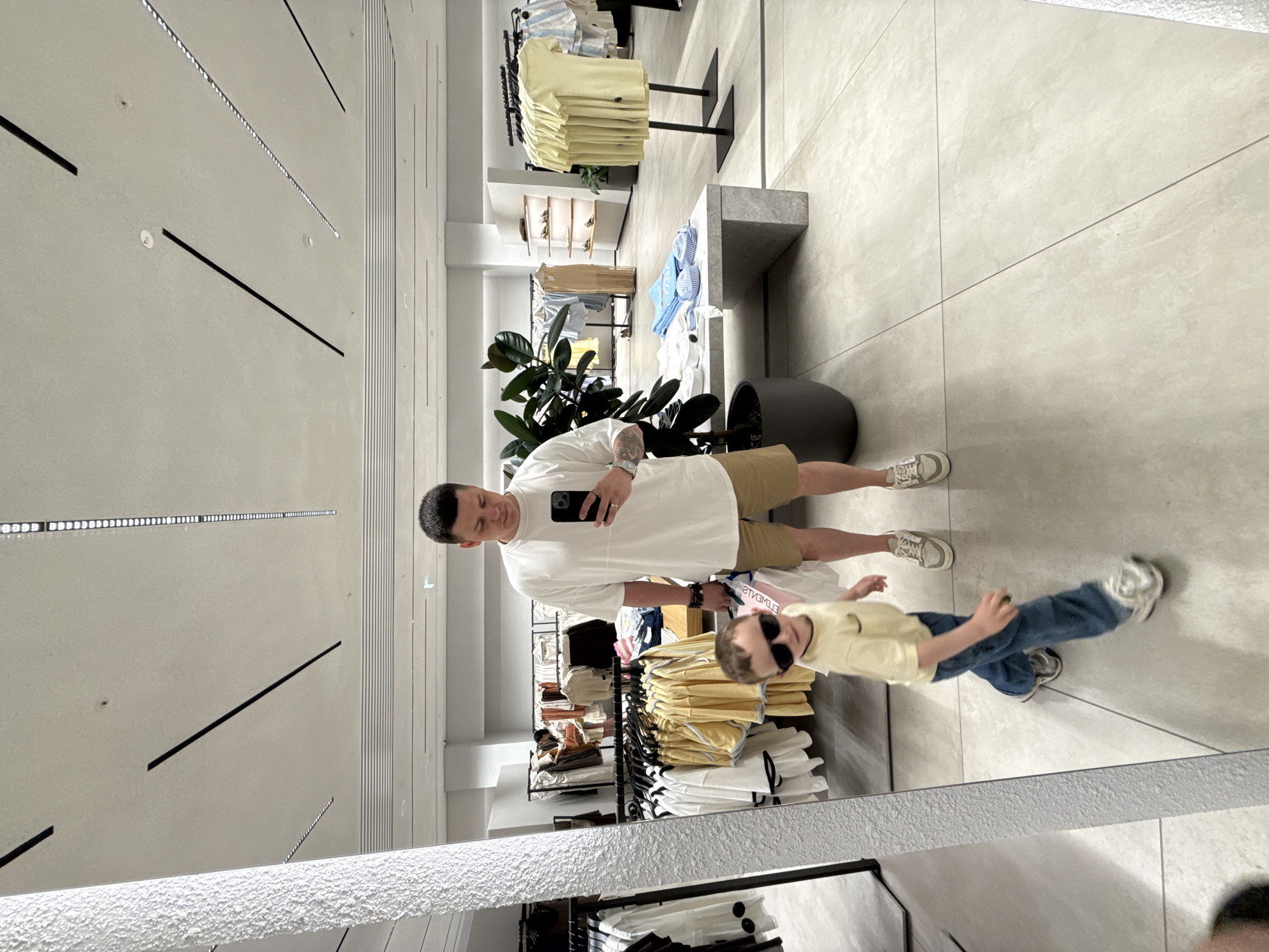 A mirror selfie in a clothing store. A man in a white T-shirt and khaki shorts holds a phone, reflecting his face. To his right, a child in a yellow shirt and blue jeans is moving. Racks of clothing, a potted plant, and neutral tile flooring are visible. The ceiling features modern lighting. The location is implied to be Vladivostok, Russia.