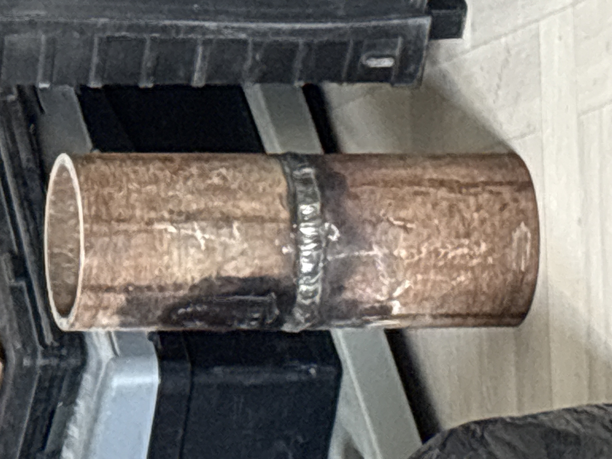 A close-up, eye-level shot shows a short, welded copper pipe standing upright on a light-colored, textured surface. The pipe has a rough, discolored surface with visible weld marks encircling it in the middle. Behind the pipe, to the left, is a dark, rectangular object with a lighter gray surface on top. To the right of the pipe is a tall, dark, textured object with horizontal ridged details. The lighting suggests an indoor environment. There are no people or animals visible in the image. The location in Zhanaozen, Kazakhstan, is not discernible from the visual information provided in the image.