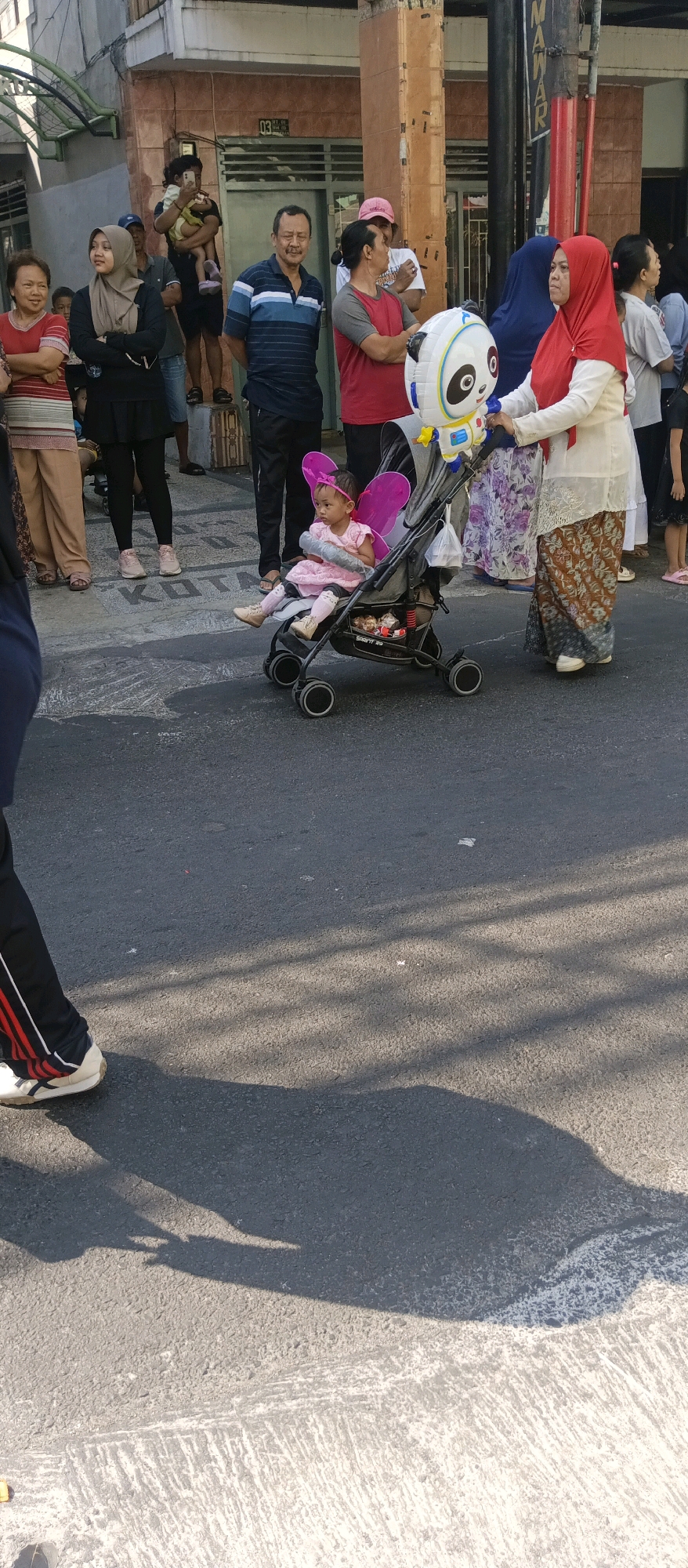 In Malang, Indonesia, a scene unfolds on a paved street, likely a public area. A young child, adorned with pink butterfly wings and a matching dress, sits in a stroller, holding a rolled-up object. The stroller is being pushed by a woman wearing a red hijab and a white top, paired with a patterned skirt. Beside her, another woman in a blue headscarf stands observing. Several other individuals are visible in the background, including a man in a striped shirt and a woman in a red shirt. The atmosphere appears to be that of an outdoor gathering or event, possibly a street festival, given the presence of a panda-themed balloon. The sun casts long shadows on the street, indicating it is daytime.