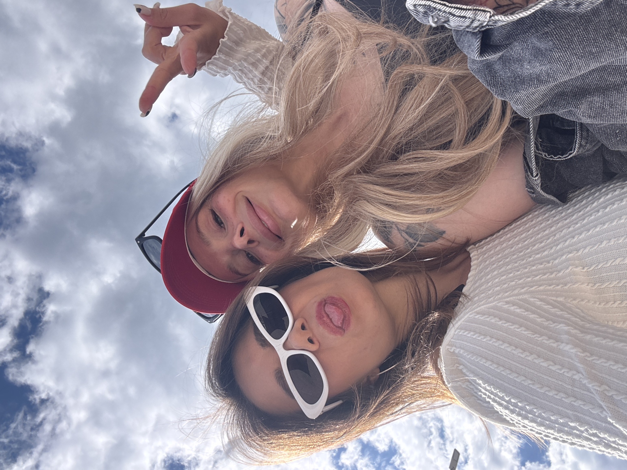 Two young women are taking a selfie outdoors, with the camera pointed up at them from a low angle. The background is a cloudy sky. The woman on the left has blond hair and is wearing a red cap, a light-colored long-sleeved shirt, and a denim jacket. She is making a hand gesture. The woman on the right has dark hair and is wearing white sunglasses and a white knitted top. She is sticking her tongue out. The location is Prijevor, Montenegro.