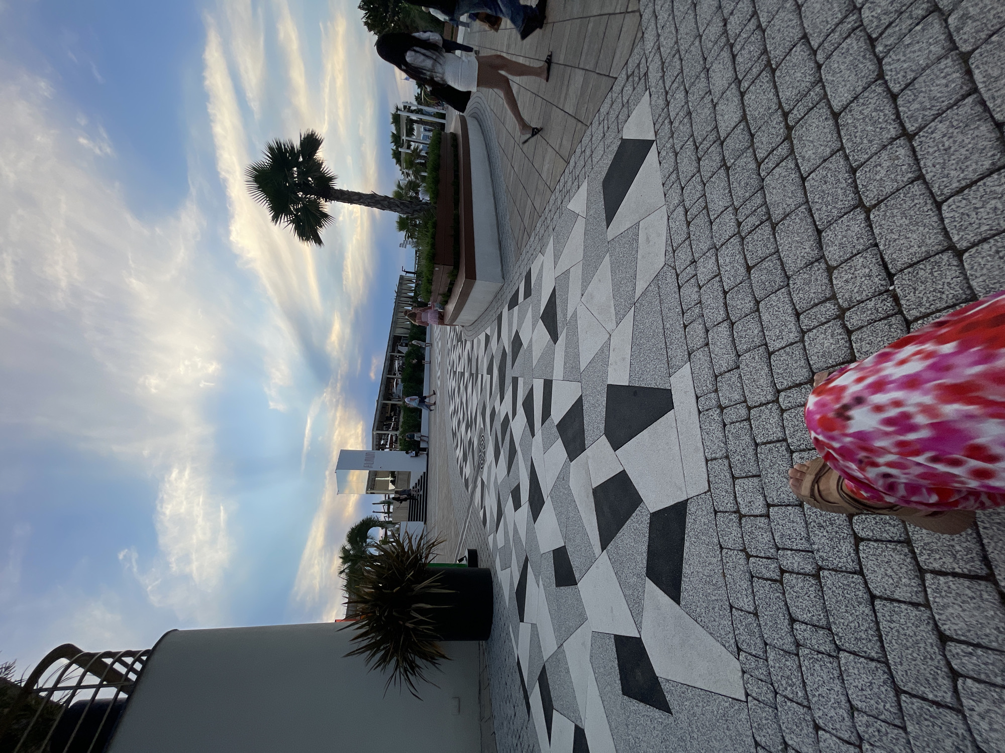The image depicts an outdoor scene in the city of Sochi, Russia, likely on a promenade or a public walkway. In the foreground, a person's legs are visible, wearing a pink and white patterned garment and sandals, sitting on a paved area composed of grey cobblestones. The walkway in front of them features a modern geometric pattern of light grey, dark grey, and black triangular and polygonal tiles.
Further into the scene, a lone palm tree stands in a raised planter box, with other palm trees and vegetation visible in the background. A white building with the word "FLAVOR" in vertical lettering on a mirrored surface is also present, suggesting a commercial establishment such as a restaurant or bar. Several individuals are walking along the pathway, some appearing to be tourists. The sky above is a blend of blue with scattered white and grey clouds, indicating either late afternoon or early evening. The overall atmosphere suggests a leisurely, urban waterfront setting.