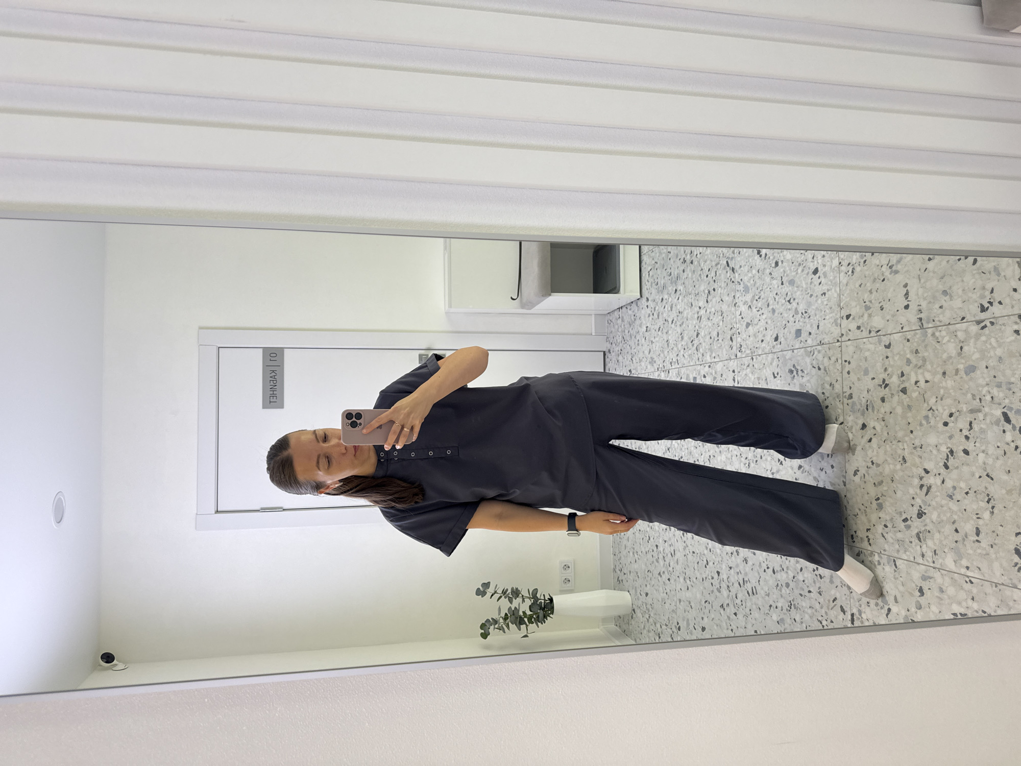 The image depicts a full-length mirror selfie taken by a woman in what appears to be a modern, clean interior. The woman, with her hair in a ponytail, is wearing a dark gray, short-sleeved collared top with button detailing and matching wide-leg pants. She is holding a pink smartphone in her right hand, taking the photo. Her left hand is visible, with rings on her fingers. She is wearing white socks.
The background shows a white wall with a security camera mounted in the upper left corner. To the right of the woman, there is a closed door with text that appears to be in Cyrillic, indicating a location within Russia. Specifically, the text on the door reads "01 КУХНЯ" which translates to "01 Kitchen". Further to the right, a white vertical blind is visible. In the lower part of the background, to the right of the woman, there is a white bench with a gray cushion, possibly a waiting area. The floor is a terrazzo-style tile with a gray and white pattern. A tall, white vase with a green plant is visible to the left of the woman, in front of the mirror.
The overall impression is one of a clean, well-lit interior, possibly a changing room, office, or a residential space. The location is identified as being in Chaikovsky, Russia, based on the Cyrillic text on the door.